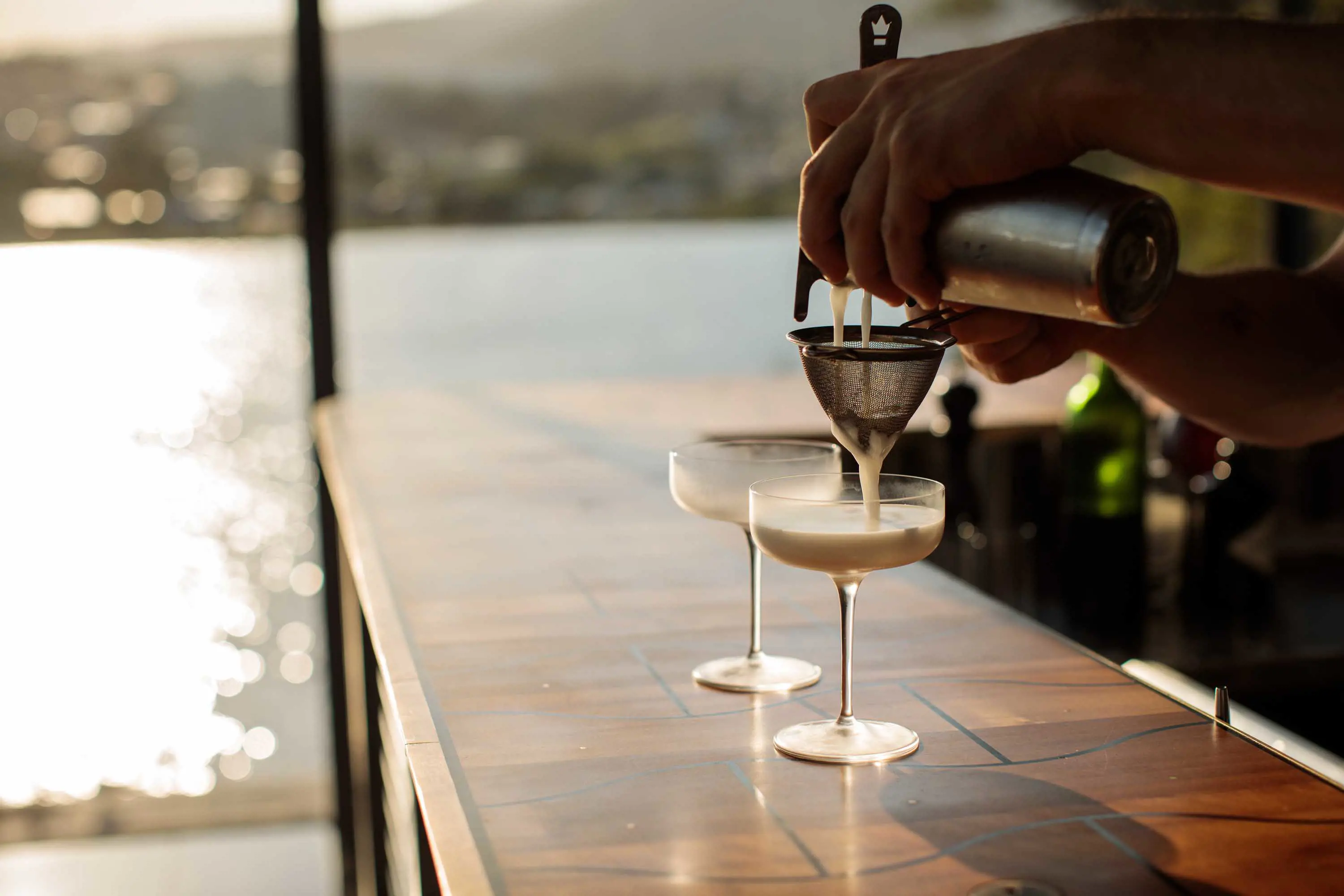 A bartender pours a cocktail through a strainer into a tall martini glass sitting on a wooden bar wit hviews of water in the background.
