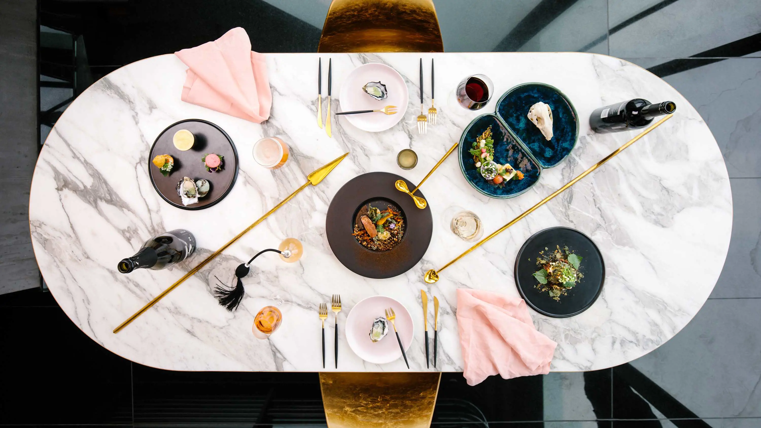 A top-down view of fine-dining cuisine presented on small plates with gold cutlery and pink fabric serviettes arranged on a marble table top.