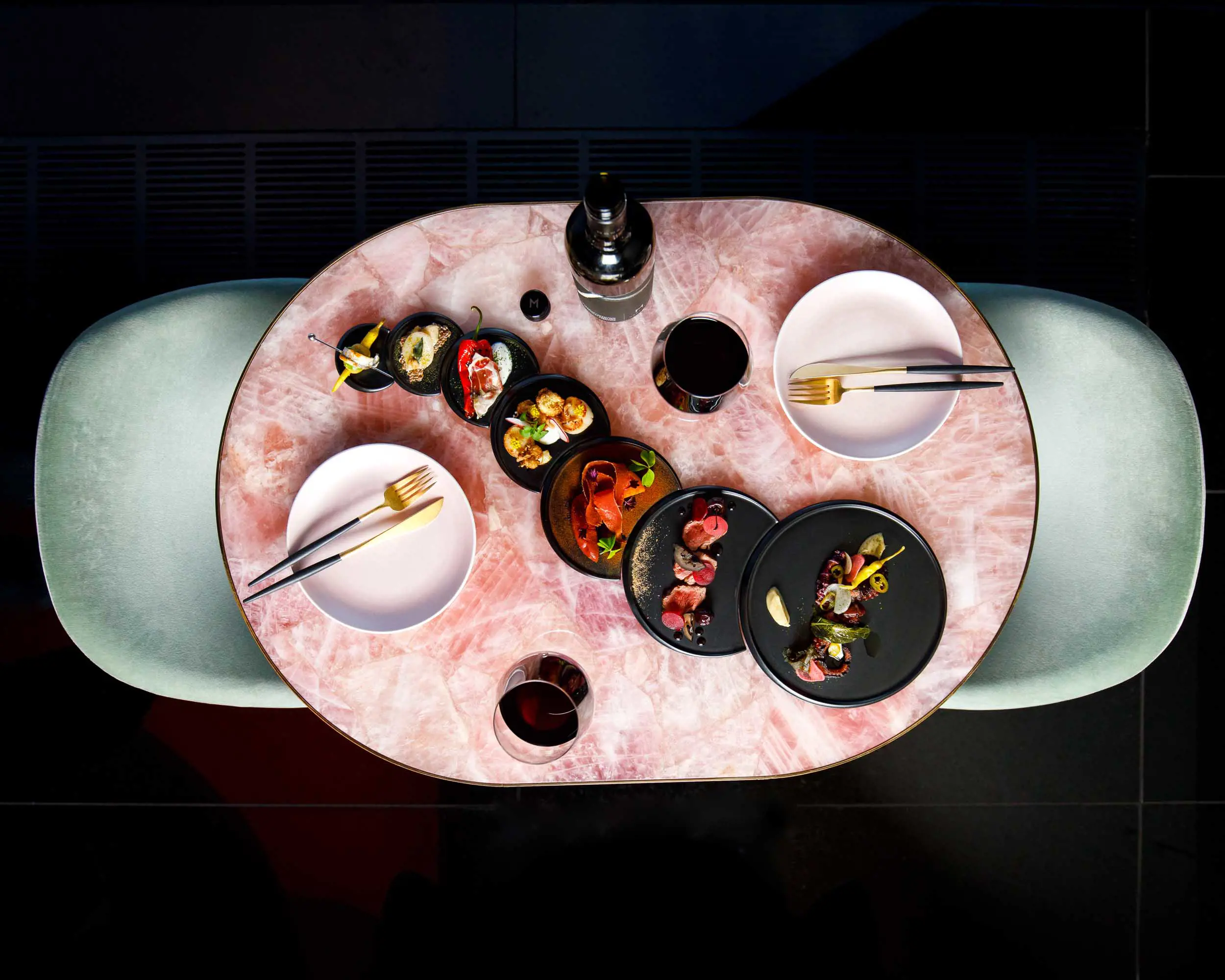 A top-down view of fine-dining cuisine presented on small plates with gold cutlery arranged on a pink marble table top.