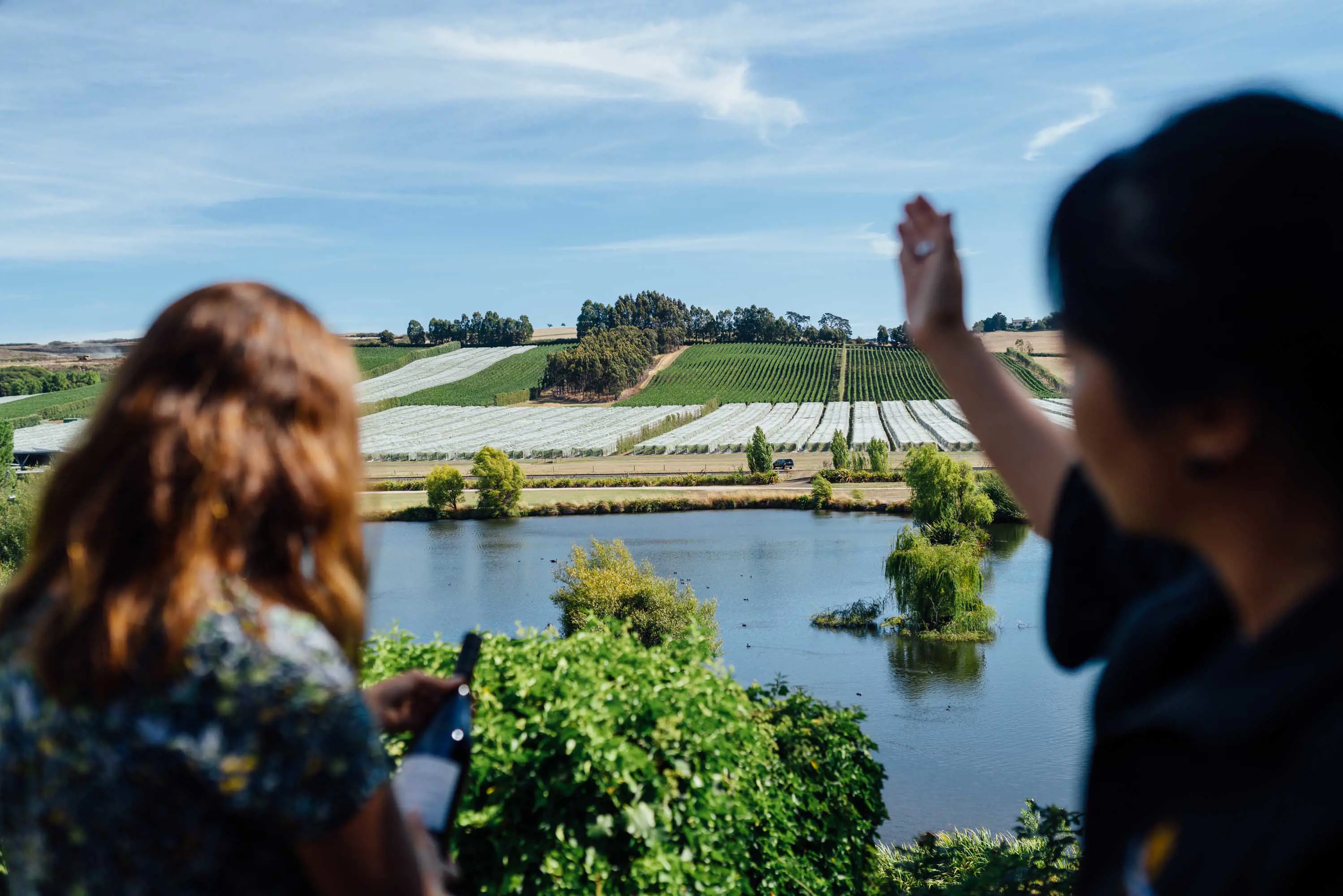 Two women stand in the foreground and look over a small lake with a vineyard stretching over hills in the distance.