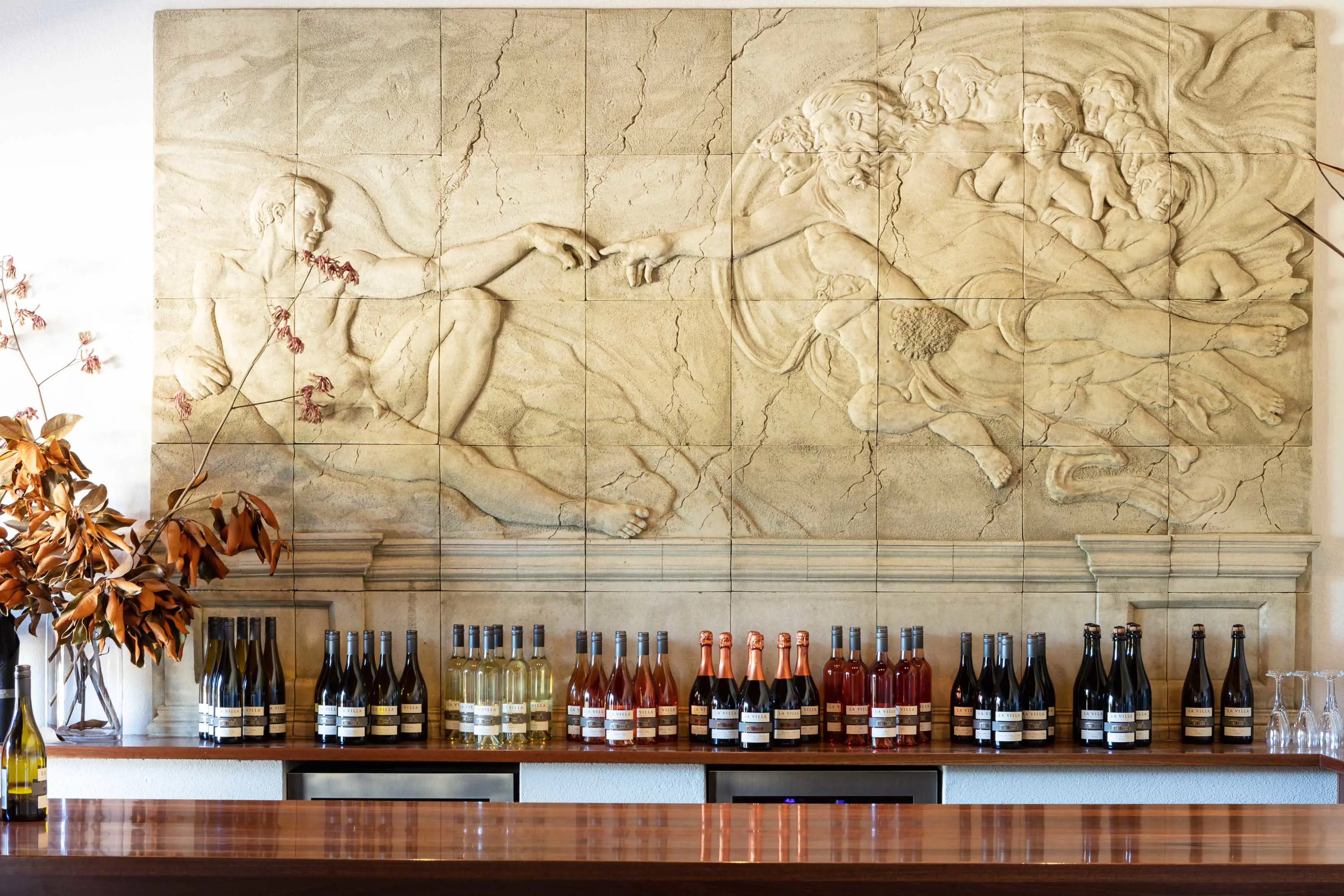 A stone relief recreation of Michelangelo's Creation of Adam is on a wall above rows of wine bottles sitting on a wooden shelf.