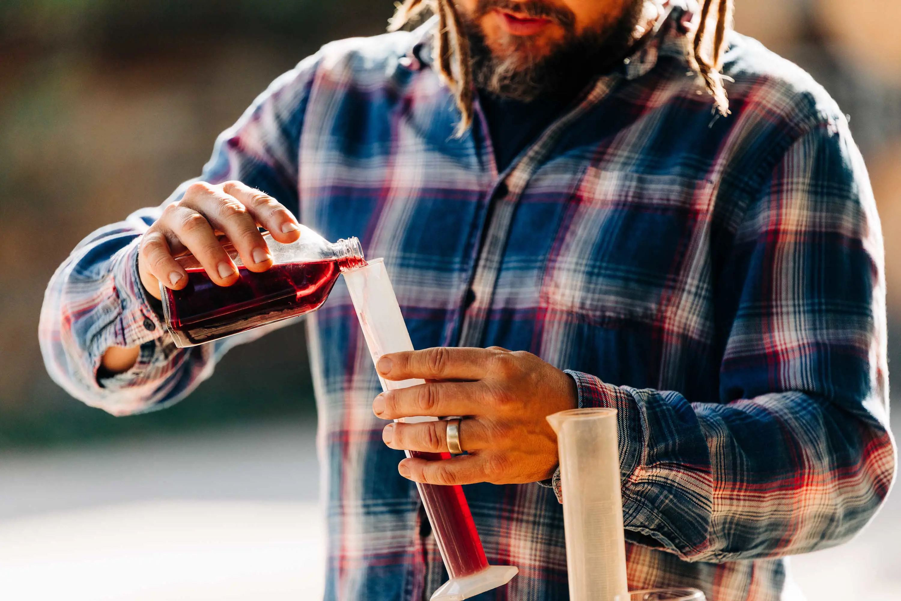 A young man with a beard, wearing a blue and red tartan shirt pours red wine from a small bottle into a measuring pitcher.  