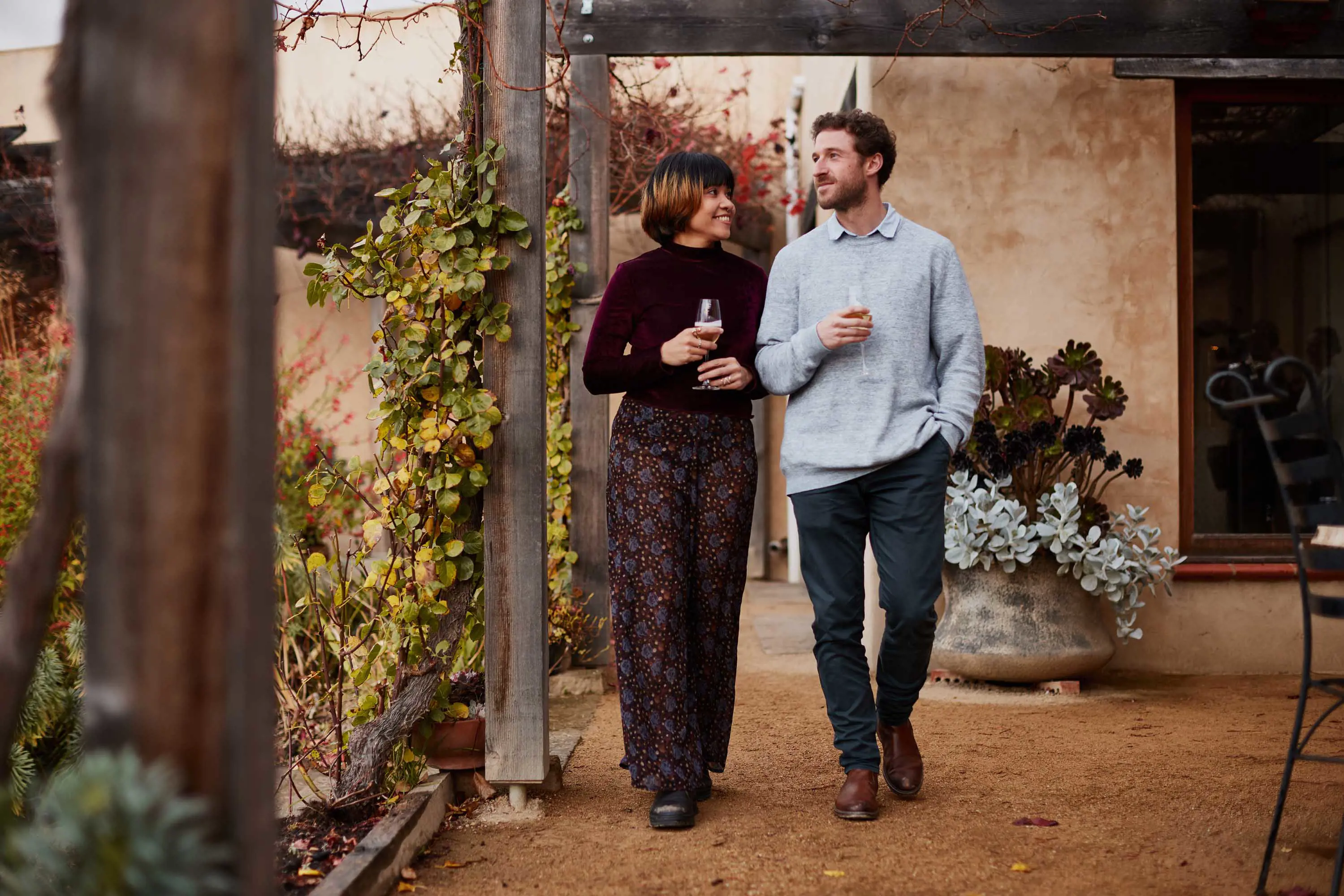 A young couple holding wine glasses and talking, walk beneath the verandah in a garden.