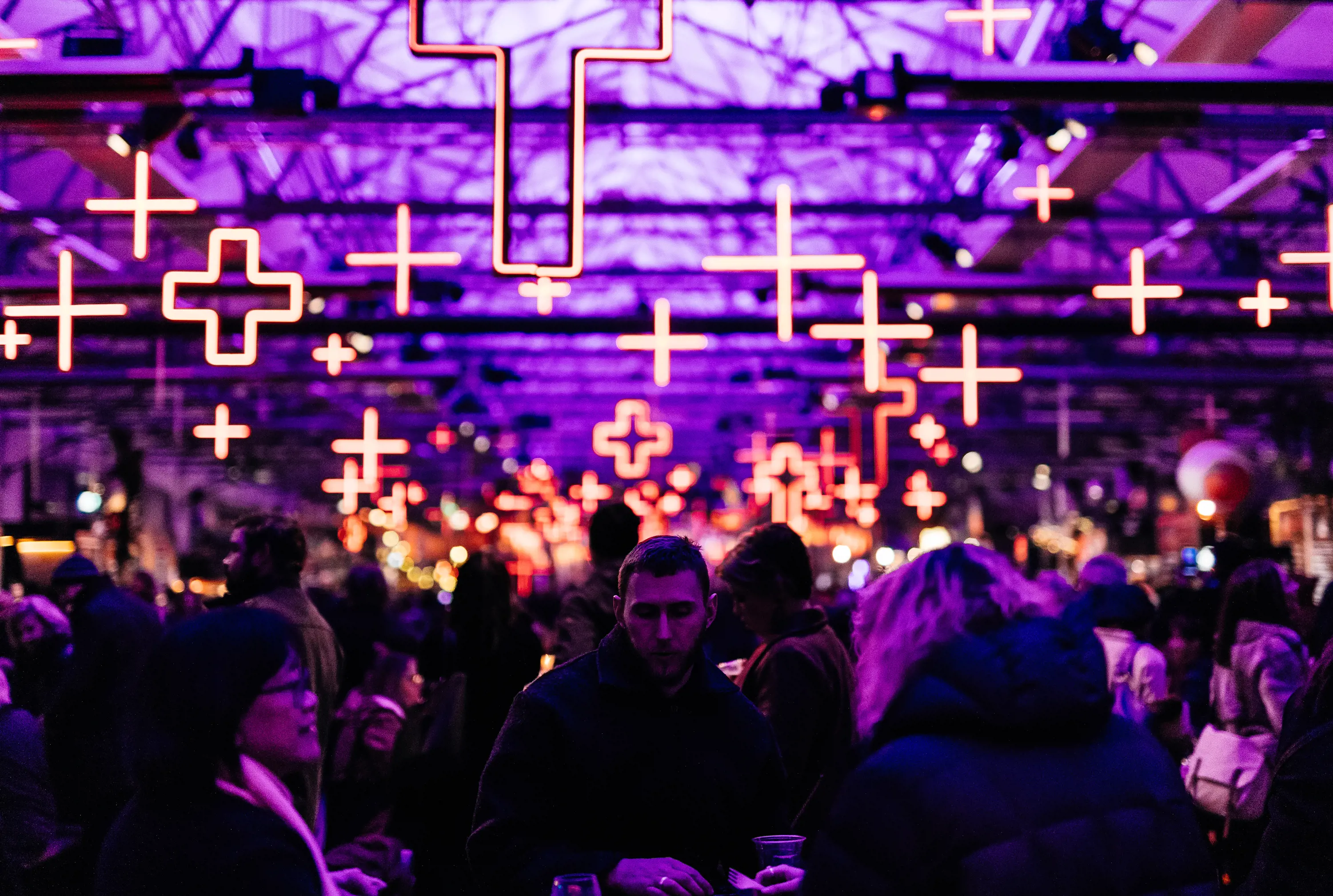 A people sit at tables at eat food under cross-shaped neon lights hanging from the rafters of an enormous wharf shed.