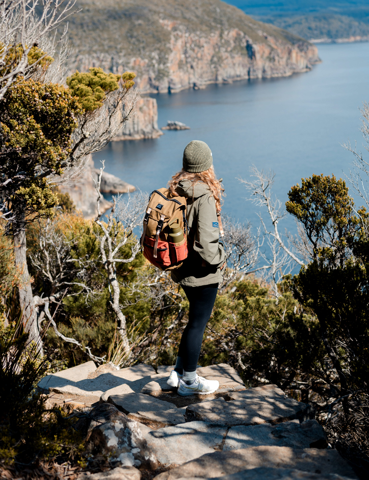 A woman wering a jacket and beanie stands in a clearing on a rocky pathway and looks over ocean and sea cliffs on a sunny day.