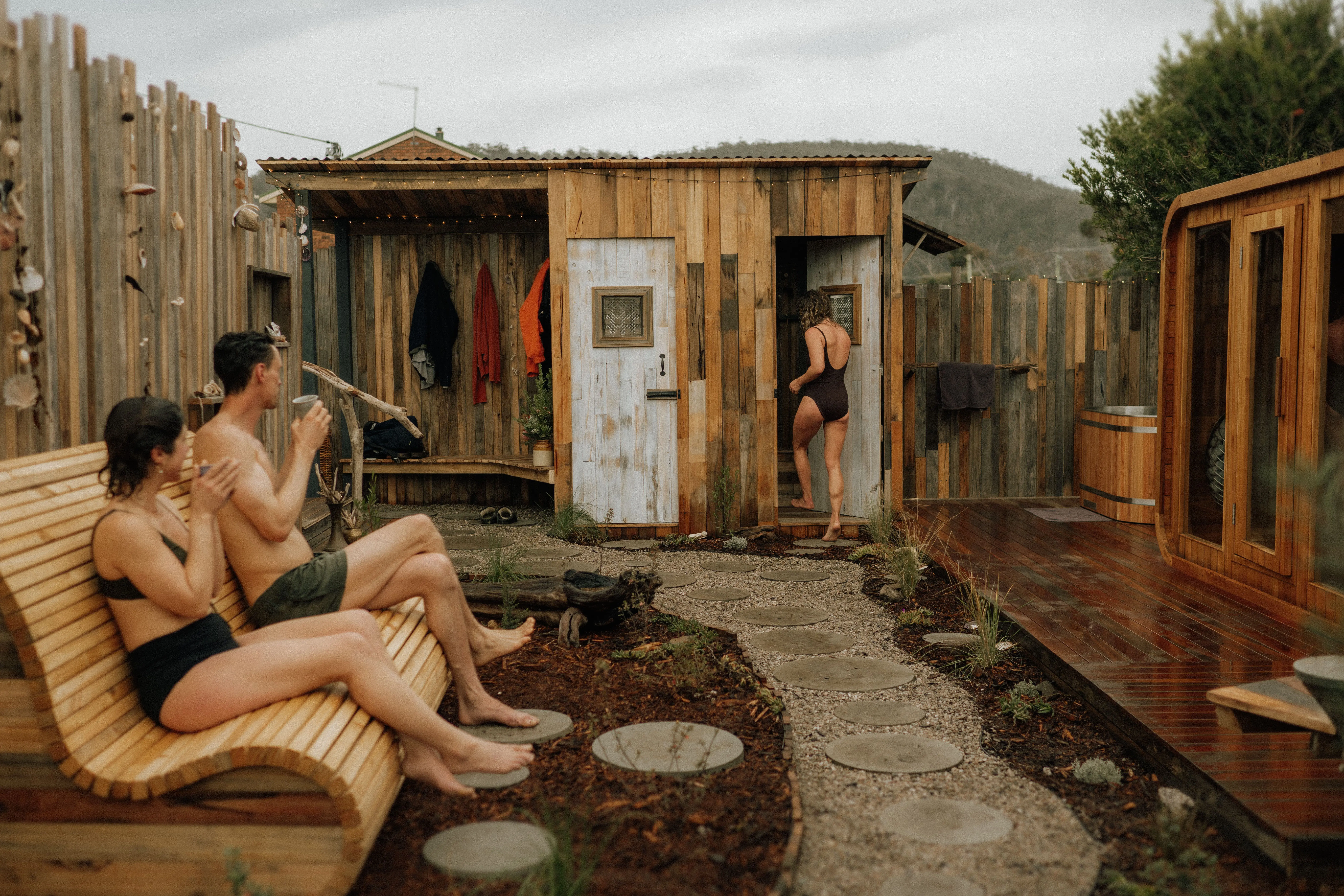 A man and woman drink from cups on a wooden bench in a garden with a stone path and timber sauna and plunge pool, surrounded by tall timber fences. A woman in bathers enters a bathhouse.
