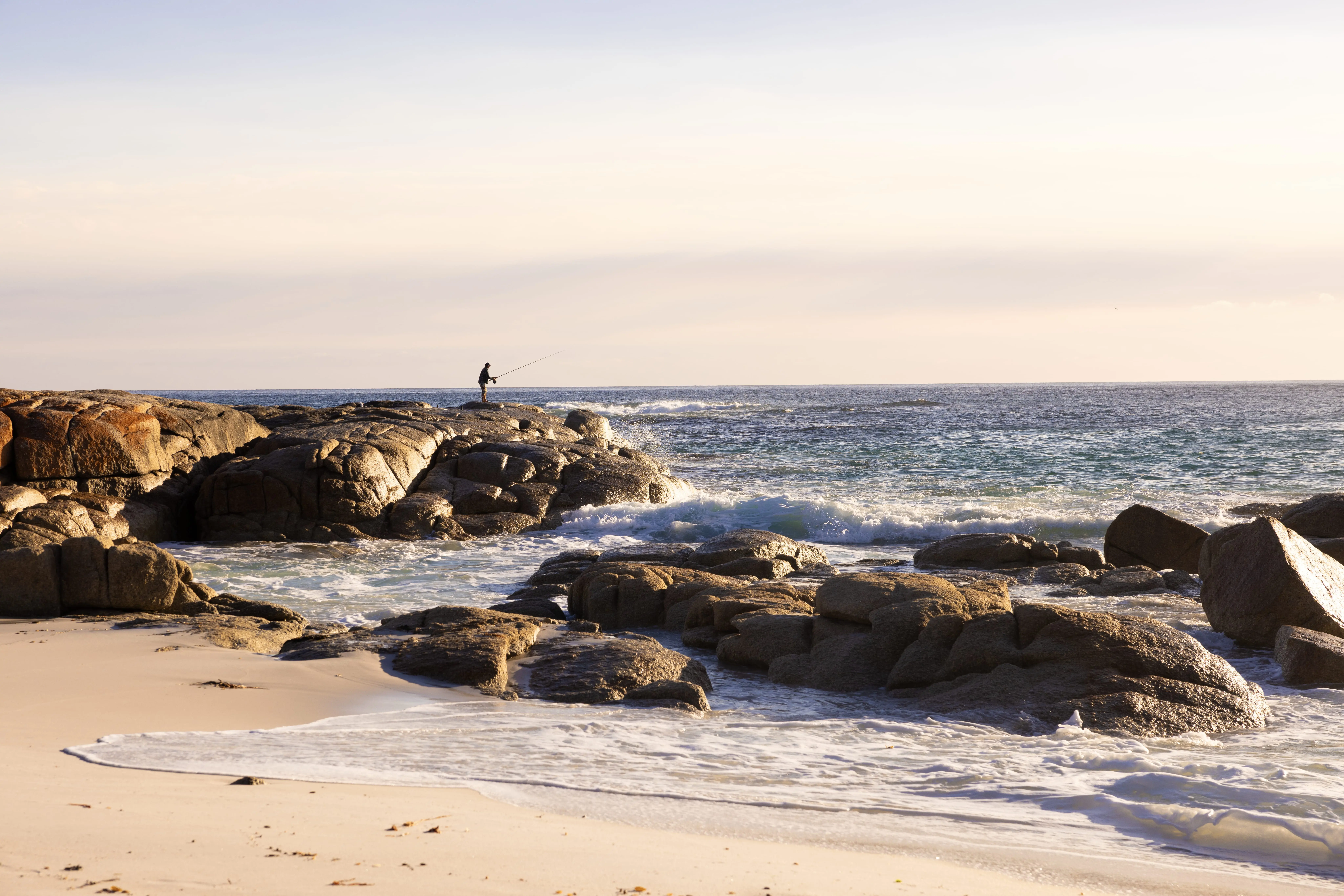 A beach scene with sand, gentle waves and various rocks and boulders. In the distance, a man silhouetted against the horizon goes fishing off the rocks.