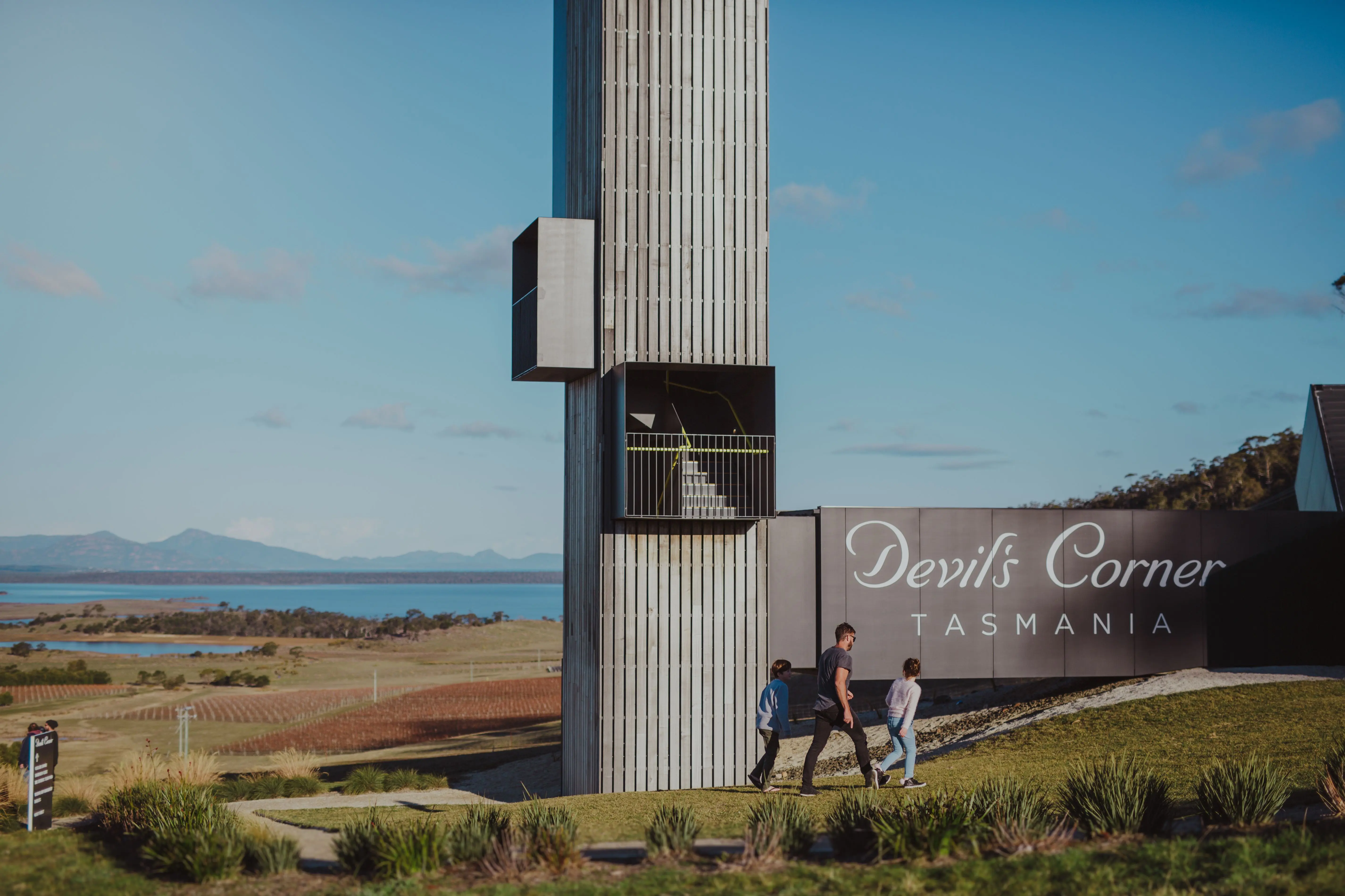 A sunny, blue-sky backdrop shows a distant blue lagoon and sea and sweeping green plains beneath mountain peaks. In the foreground, a man and two boys walk towards a structure that reads "Devil's Corner Tasmania". The modern design timber tower is narrow, tall and rectangular with a staircase within.