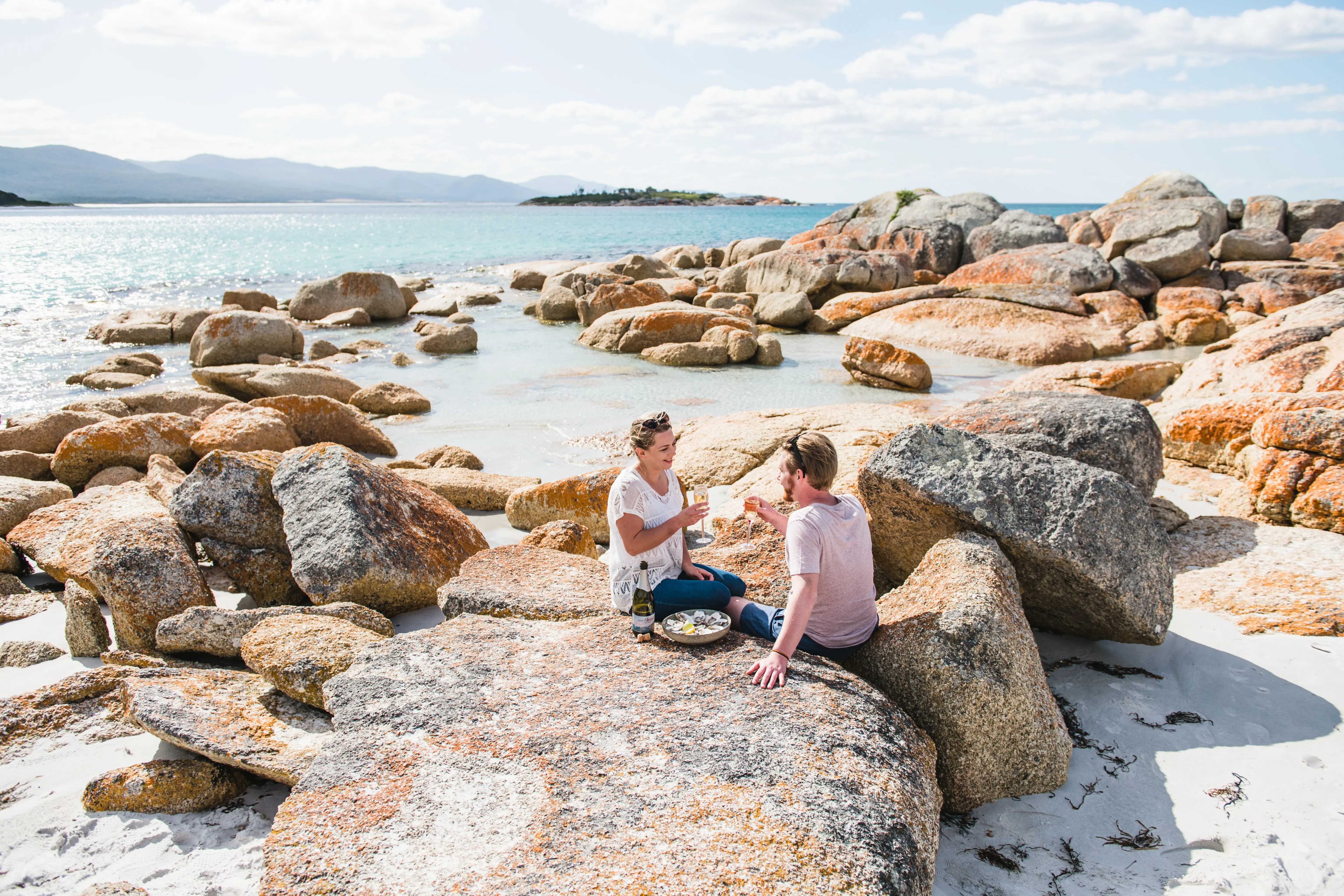 A man and woman wearing white T-shirts cheers two glasses of wine while sitting on a boulder, at a beach strewn with various orange, yellow and grey rocks. I the distance behind them is a blue sea and the green Diamond Island.