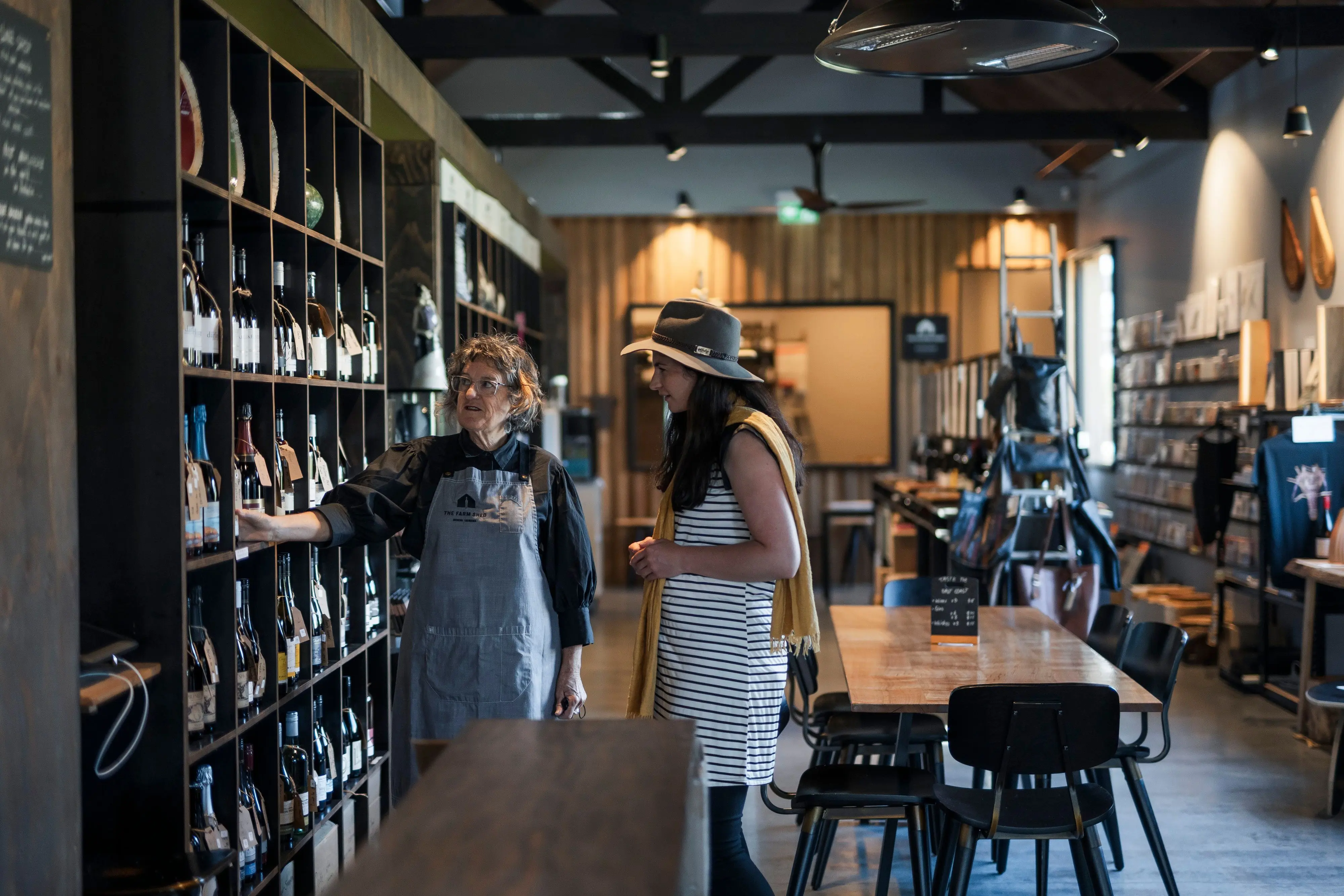 A woman in a wide-brimmed hat looks attentively at where another woman in an apron is pointing at several bottles of wine on a grid shelf. The pair are standing inside a timber shed with dining tables and chairs throughout. 