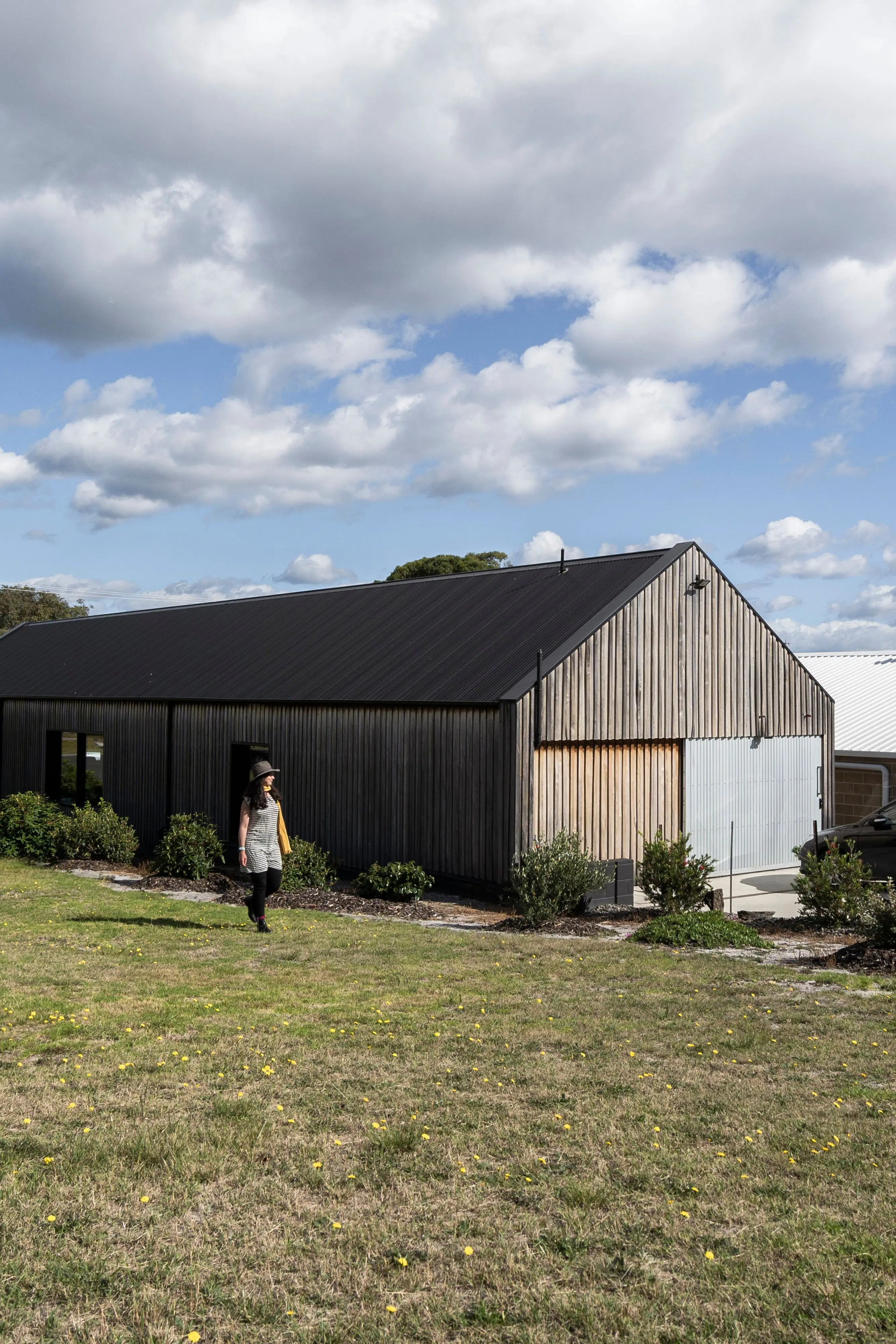 A woman in a wide-brimmed hat walks along a green lawn next to low hedges beside a long, timber gable barn in the sun.