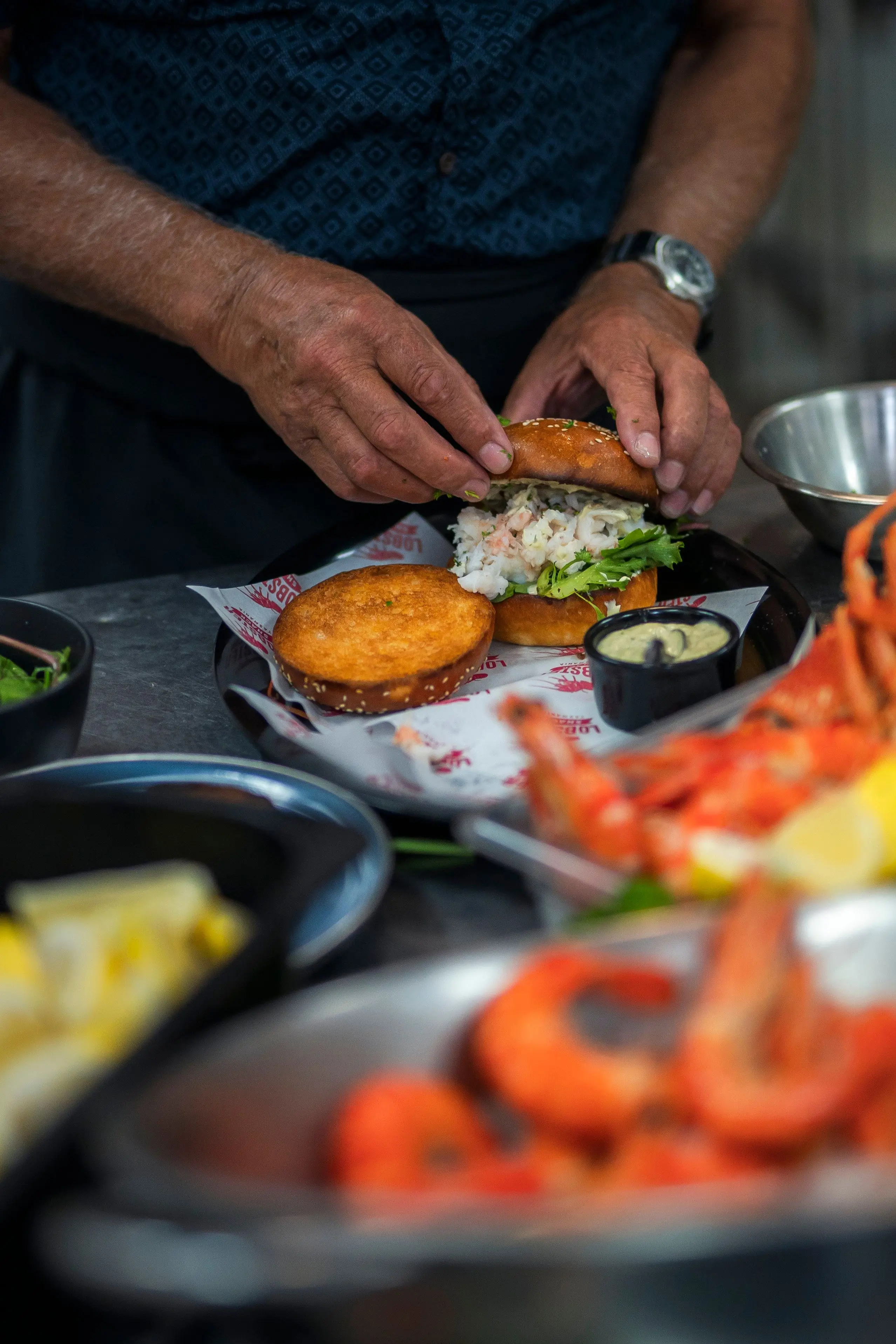 A man's hands place a golden seeded bun on top of a burger filled with fresh lobster, mayonnaise and lettuce, prepared on a plate next to creamy dipping sauce. Out of focus in the foreground is a tub of vibrant orange prawns and wedges of lemon.