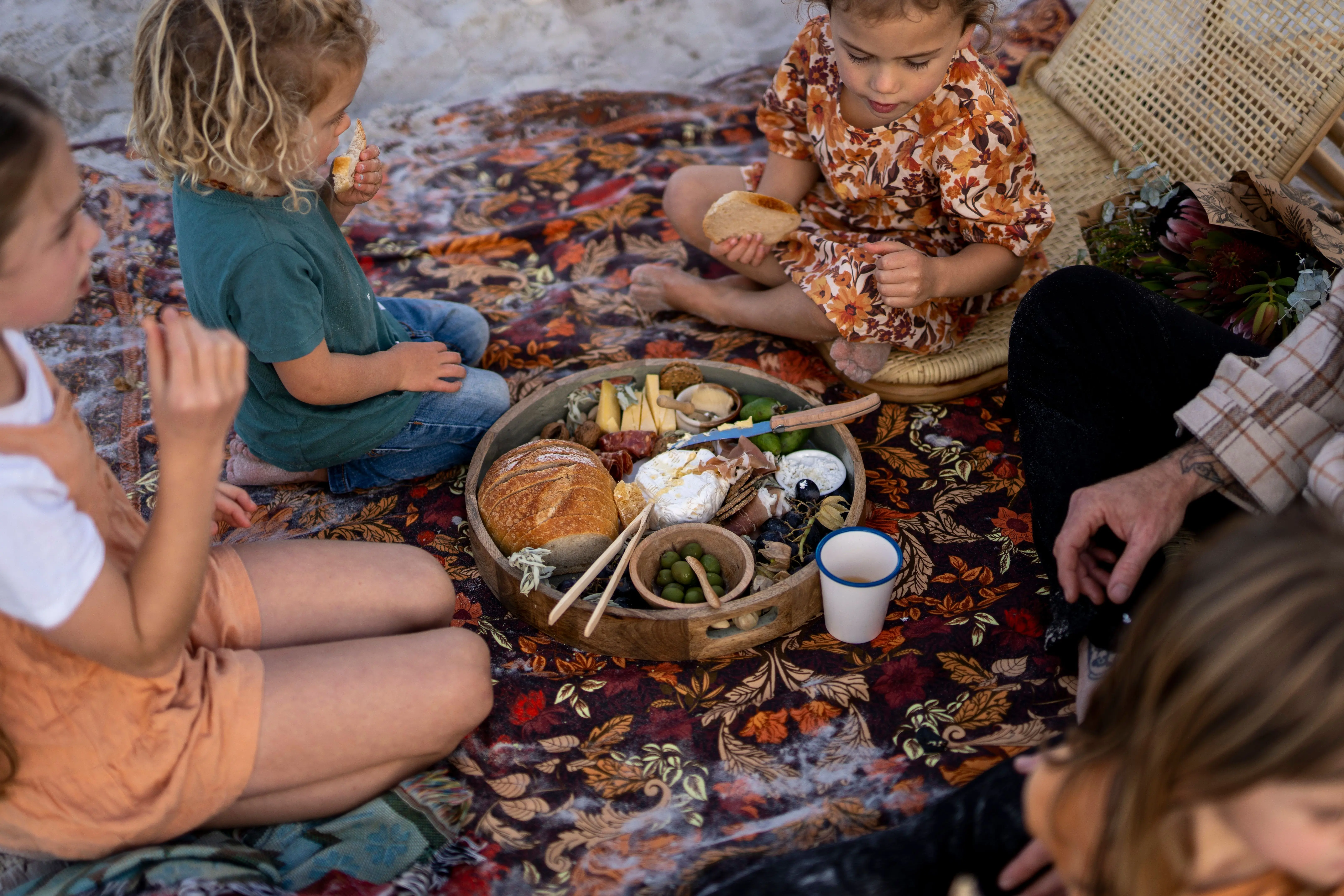 Four children and a parent sit on an ornate orange, red, gold and brown picnic rug on a beach, eating from a platter or artisan bread, meats, olives, cheeses and condiments.
