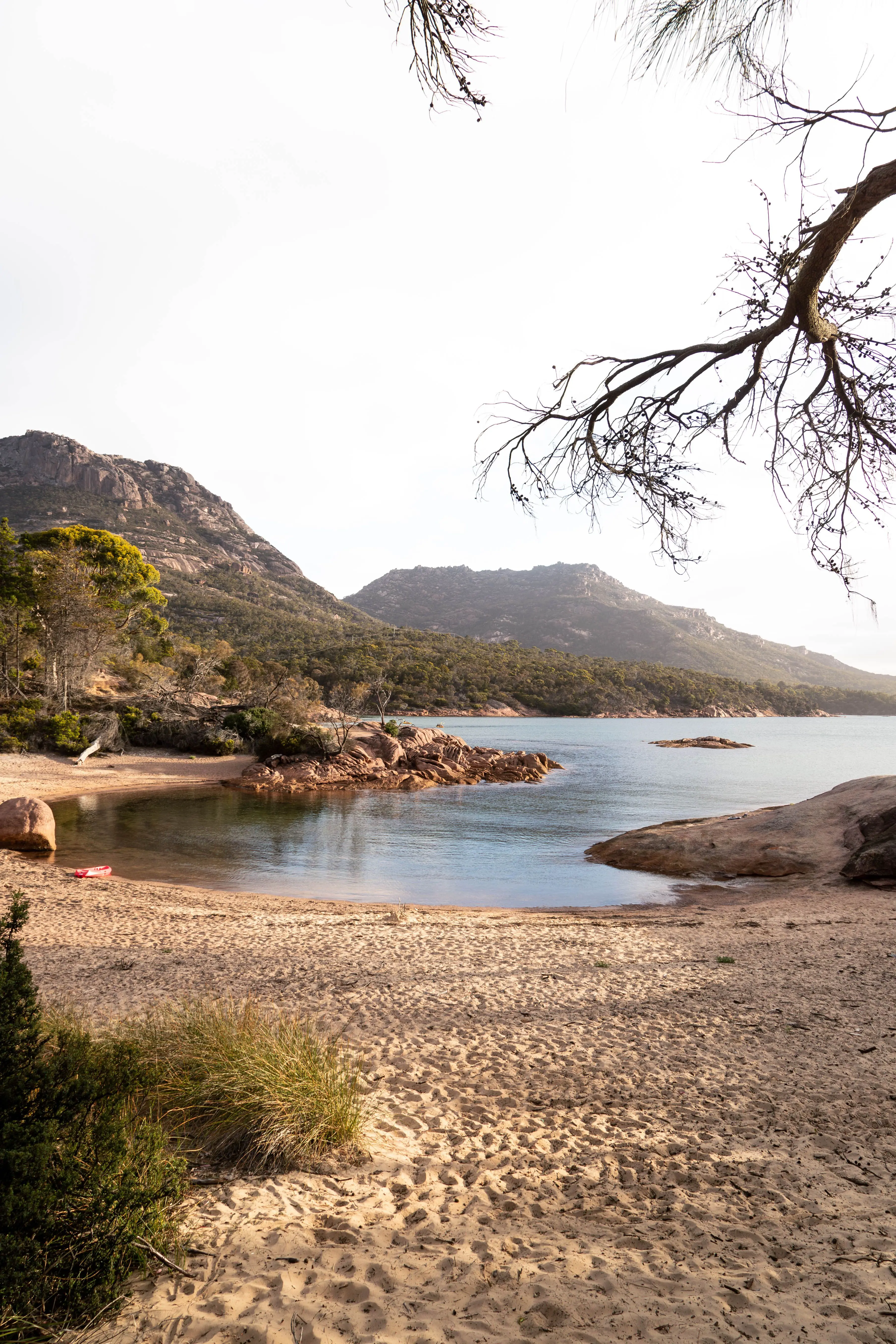 A small, glassy beach with a red kayak on the shore is surrounded by gold sand, sloping rocks and eucalypt trees in the afternoon sun. In the distance, two steep pink-granite peaked mountains covered in trees loom.