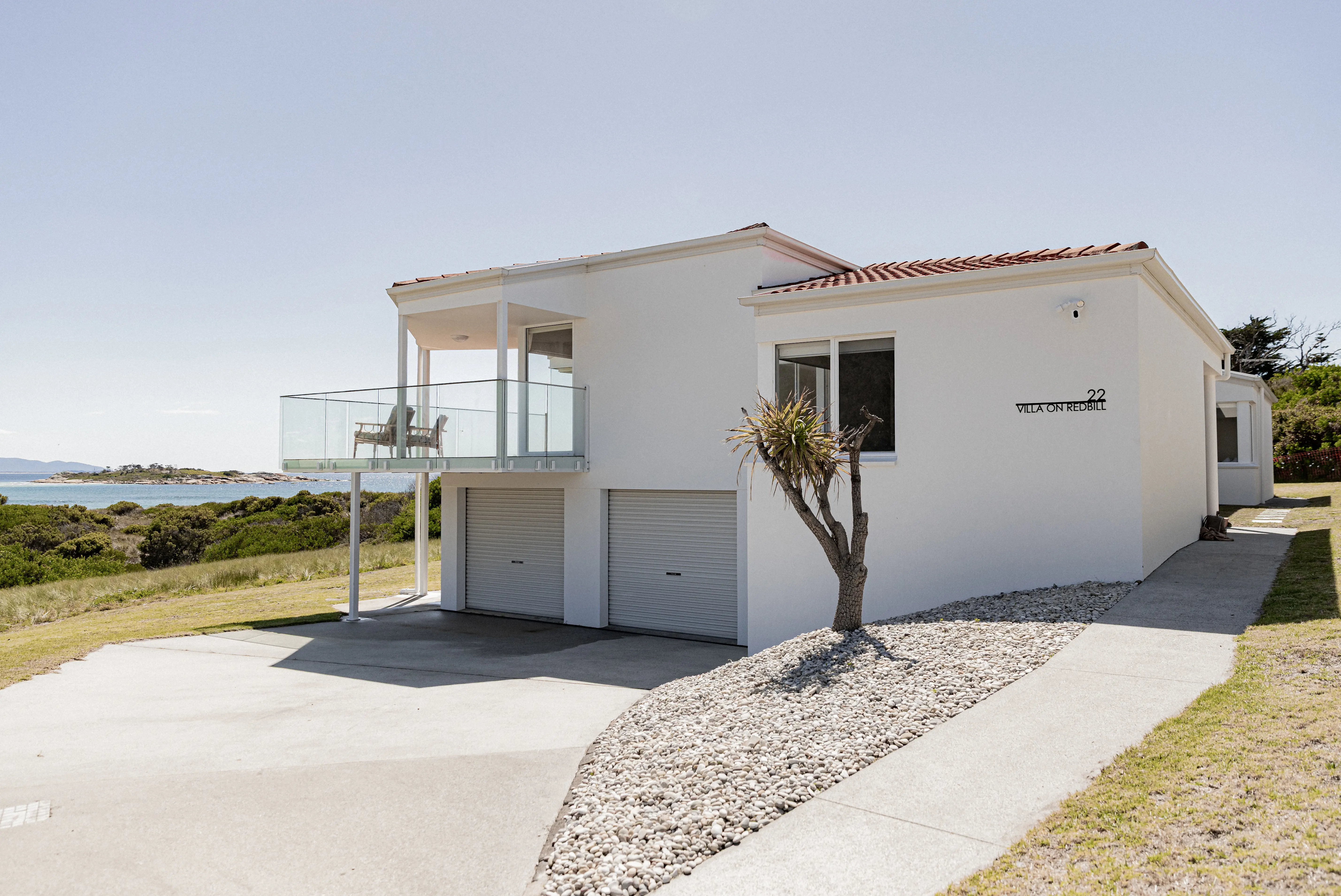 Modern white coastal home with a glass-fronted balcony, double garage, and landscaped driveway, set on a grassy block with ocean views of Diamond Island in the background.
