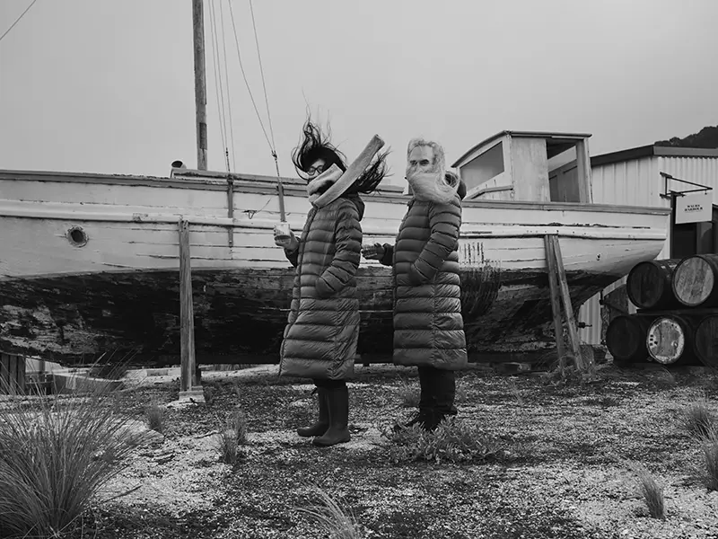 Two people wearing body-length puffer jackets stand in front of a large wooden boat up on stilts, their scarves and hair blowing wildly in the wind.
