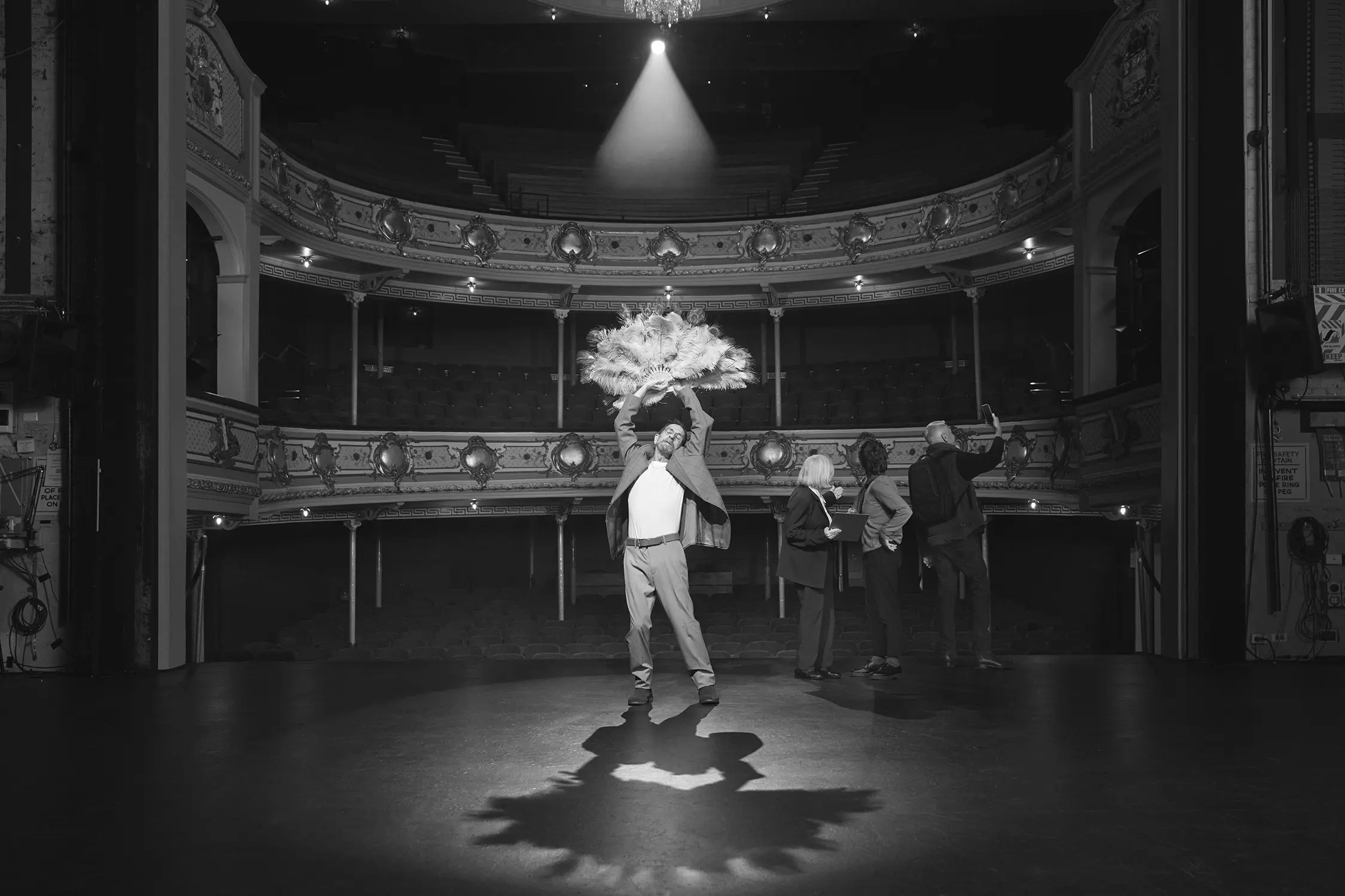 A man dramatically poses with under the spotlight on a stage. People on a tour of the historic theatre ignore him, taking photos of the space.