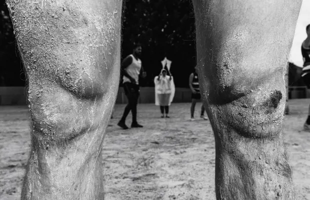 A close-up of human knees caked with dried mud. In the background, a group of people blurred in motion, in the middle of a football game.