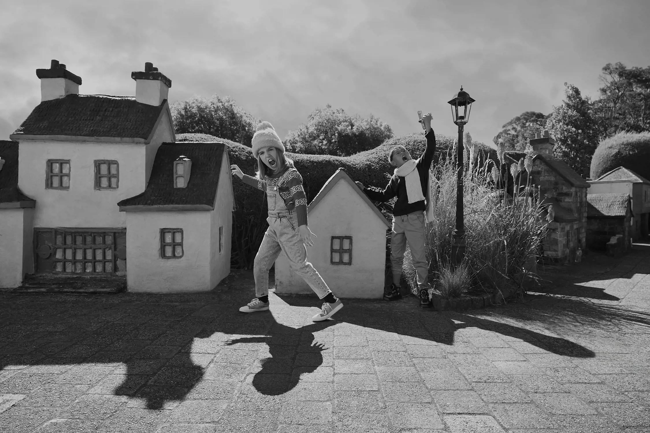 Two kids dressed warmly in scarves and beanies roar and playfully stomp through the streets of a miniature village.