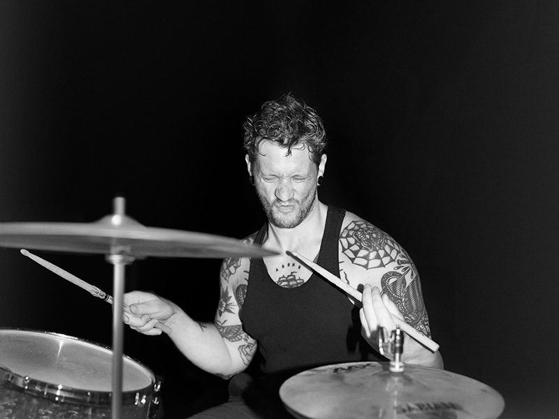 A man wearing a black singlet with heavily tattooed arms plays a drumkit, his face scrunched up mid-headbang.