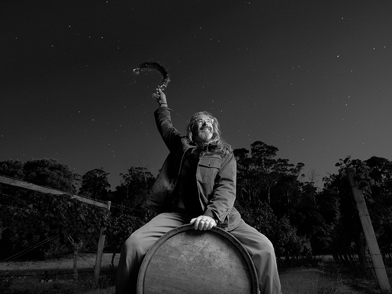 In a paddock under the night sky, a man straddles a fallen barrel, holding on with one hand. With the other, he throws the contents of a wine glass into the air.