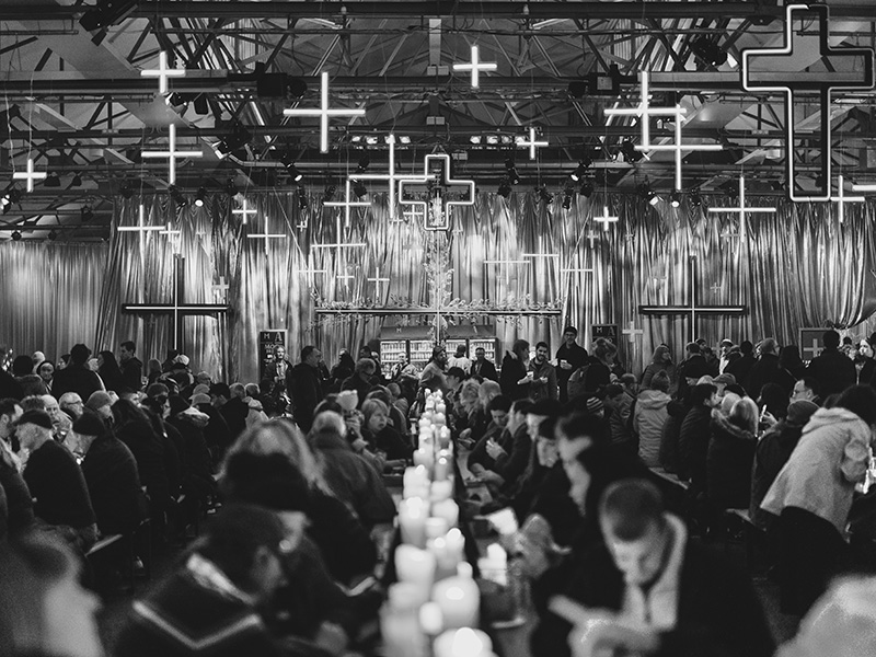A crowded venue filled with people, illuminated by glowing crosses hanging from the ceiling and lined with candles on the tables.