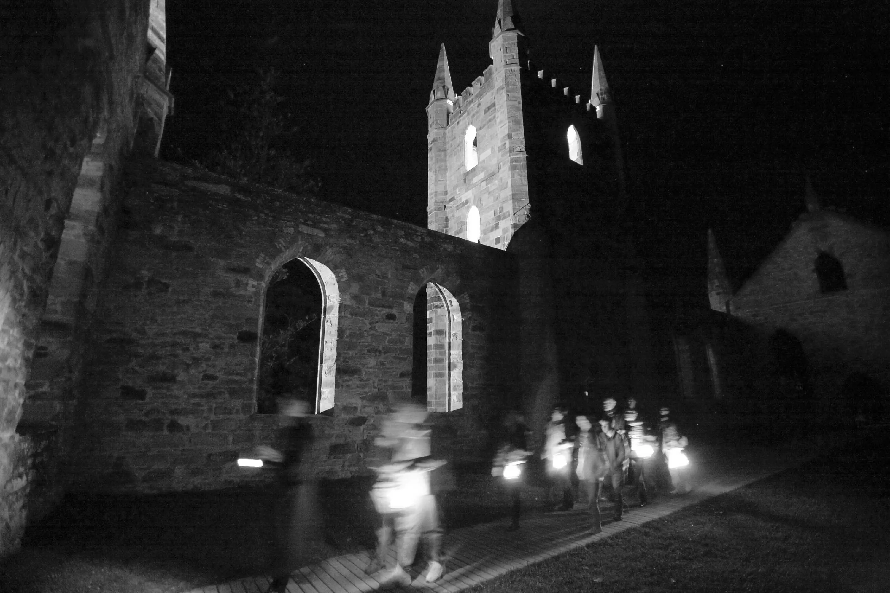 A tour group holding torches walks along a path in front of a colonial era church spire made from sandstone is illuminated by lights from below.