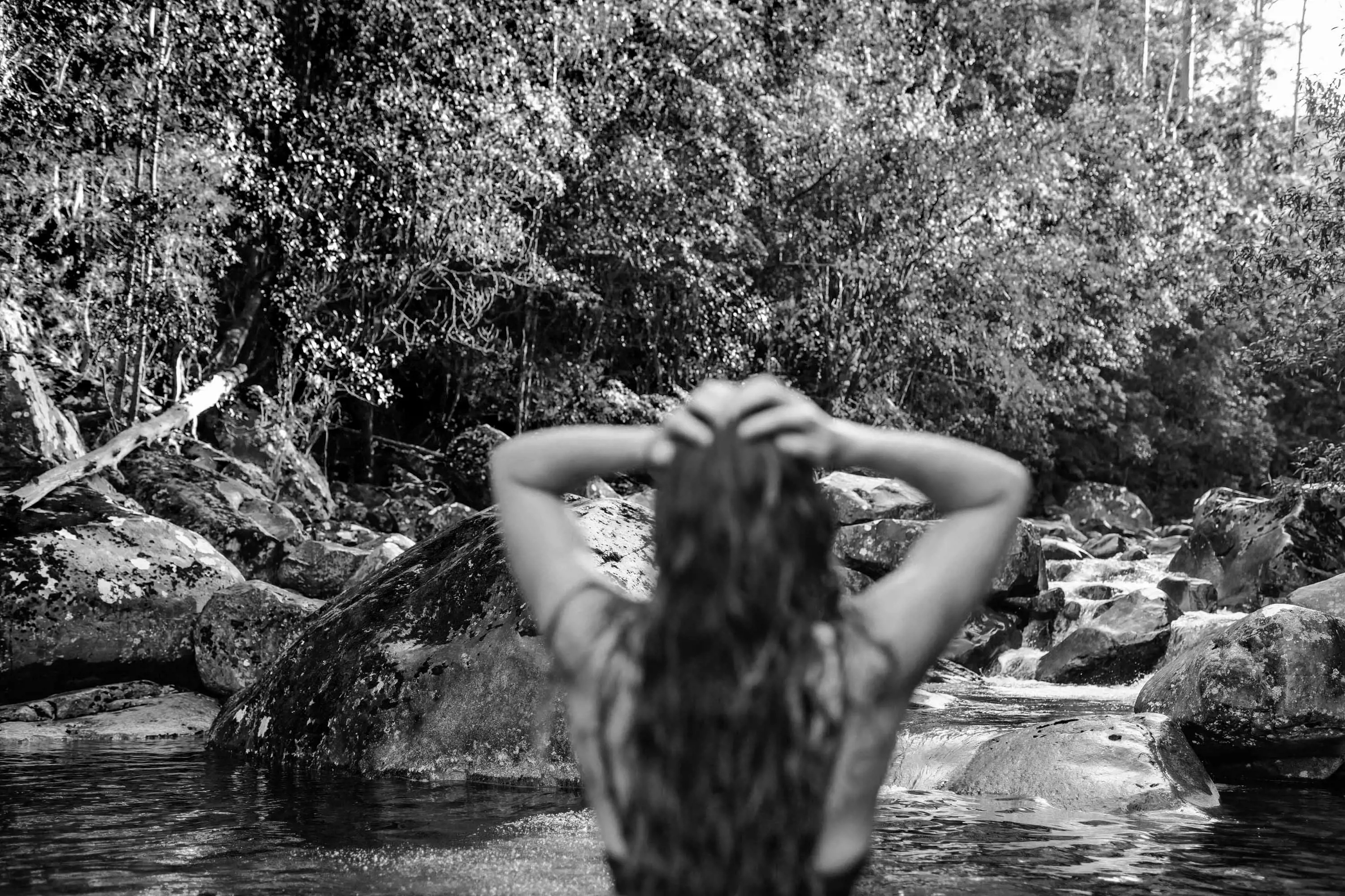 A young woman stands in rocky waterhole within a dense forest.