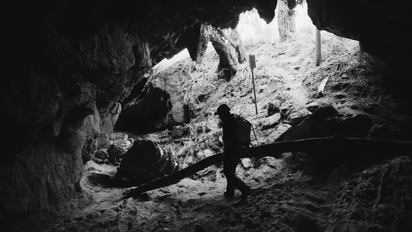 A woman in protective helmet and clothing walks carefully into the base of a dusty cavern. Light illuminates the base from a large hole above.