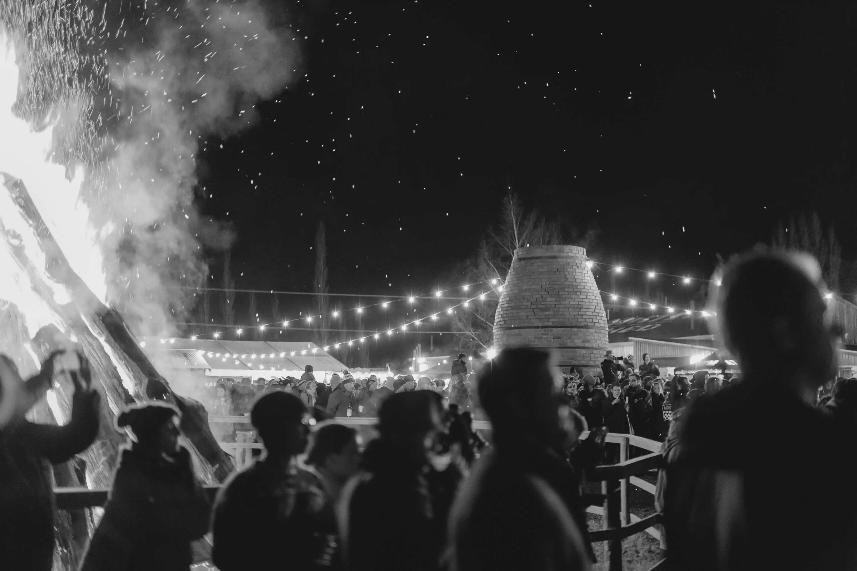 A large group of people gather under lights strung overhead and next to a large bonfire, that billows sparks and smoke into the dark night air.