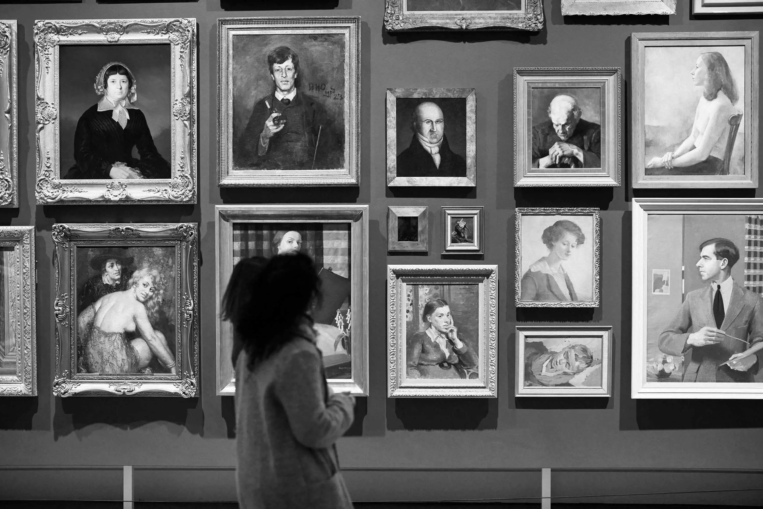 A woman walks by a tall wall with an arrangement of portrait paintings in various sizes with lavish frames. 