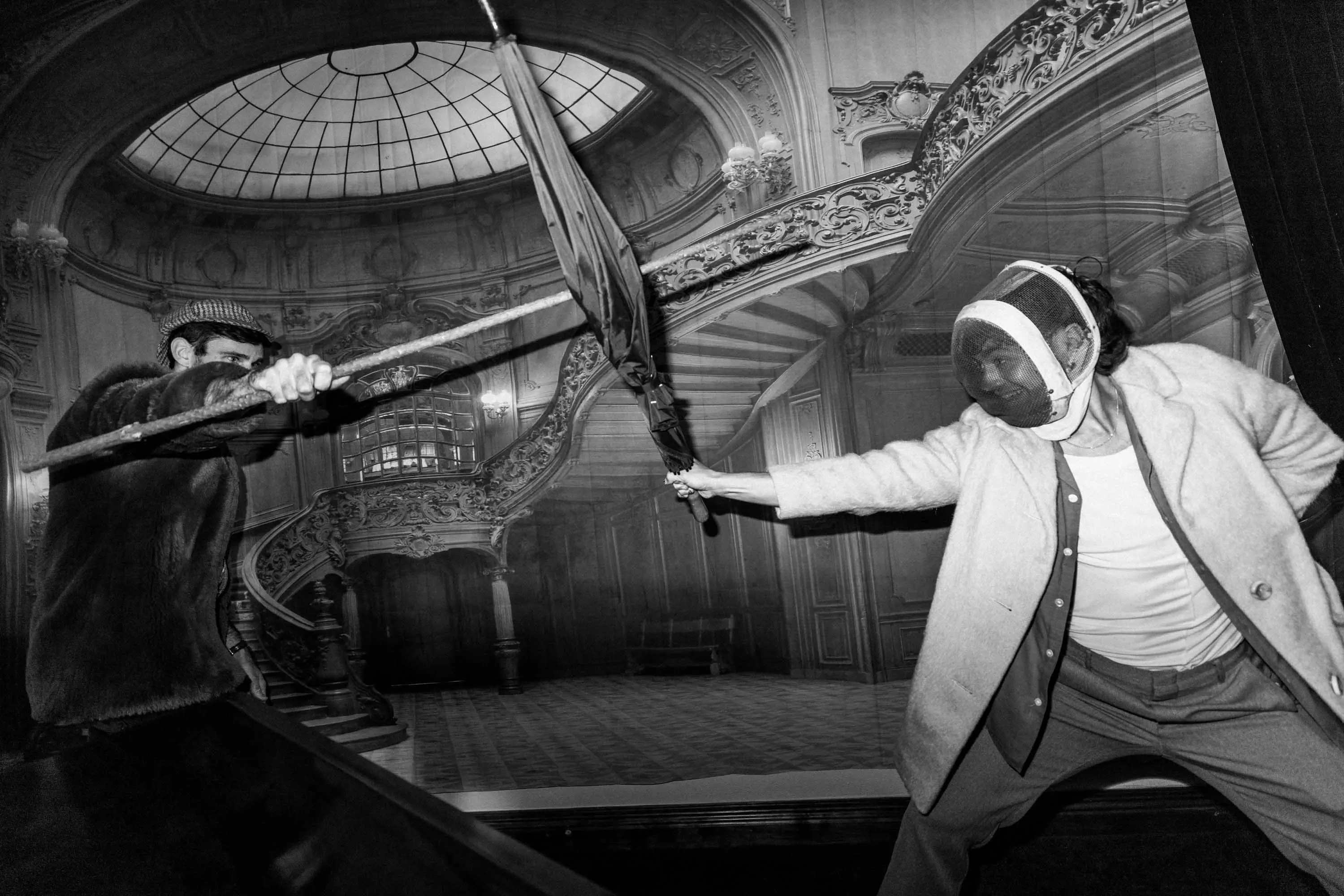 Two men, one wearing a fencing mask, mock-sword fight with an umbrella and a stick in from of photographic wallpaper depicting a large hall.