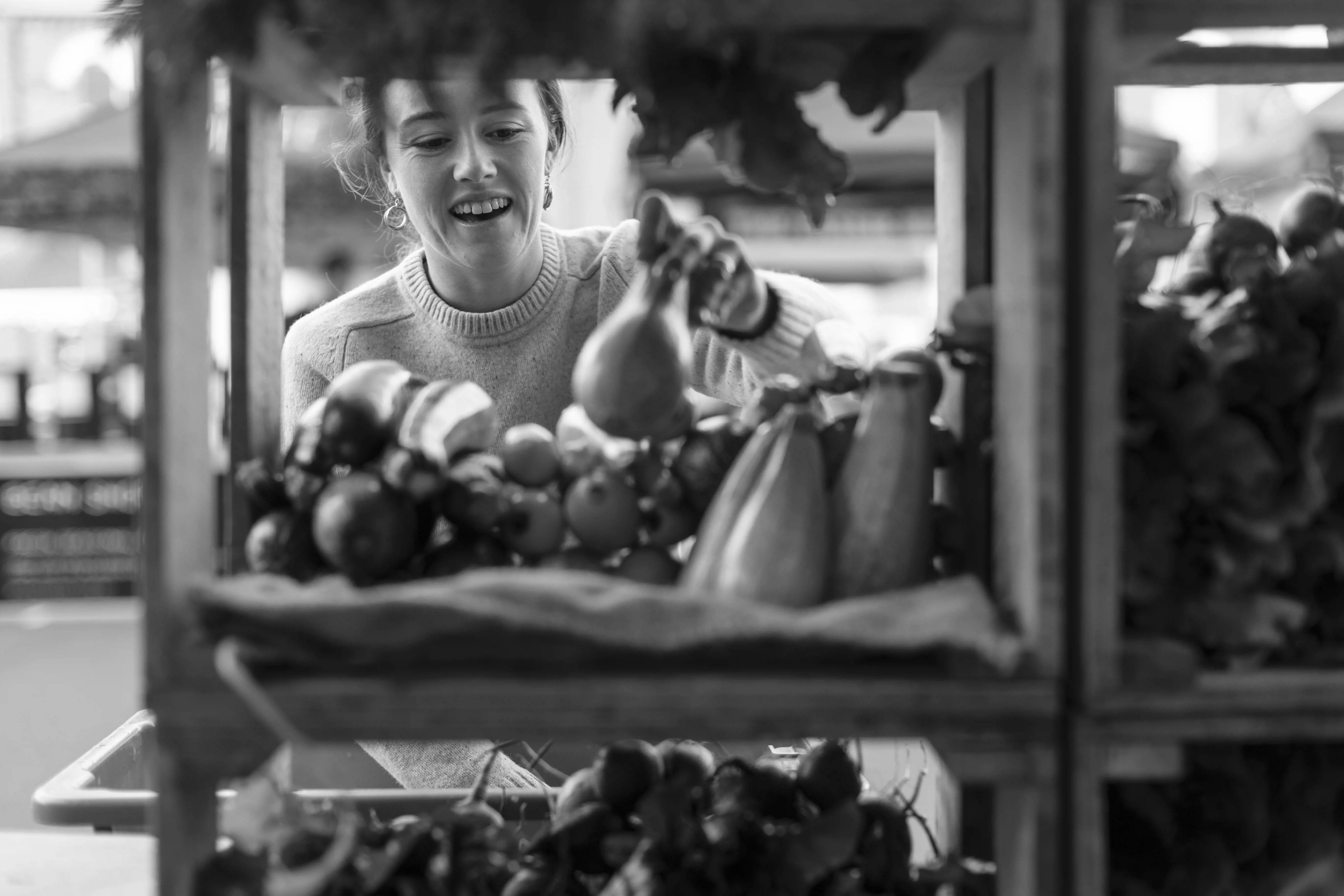 A young woman picks from vegetables displayed in wooden shelves at a market stall. 