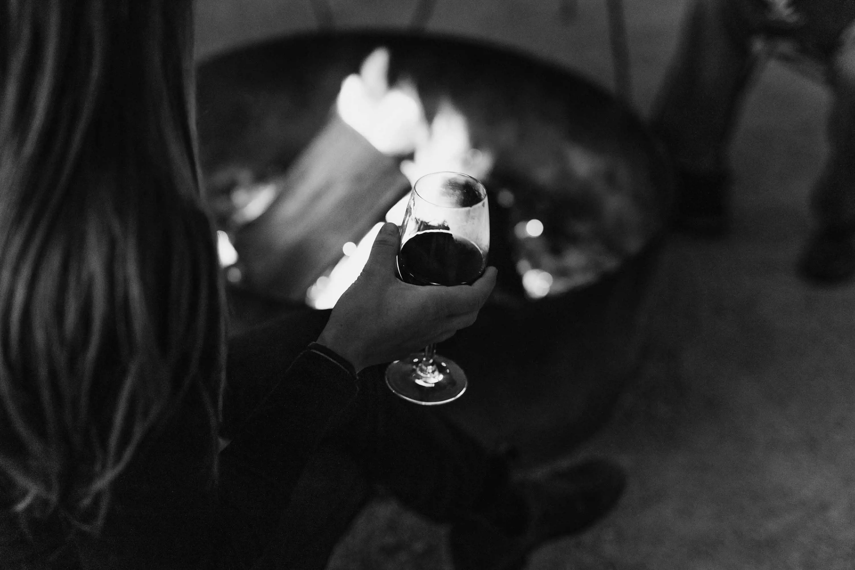 A woman sits beside a fire pit, reading a book and drinking red wine.