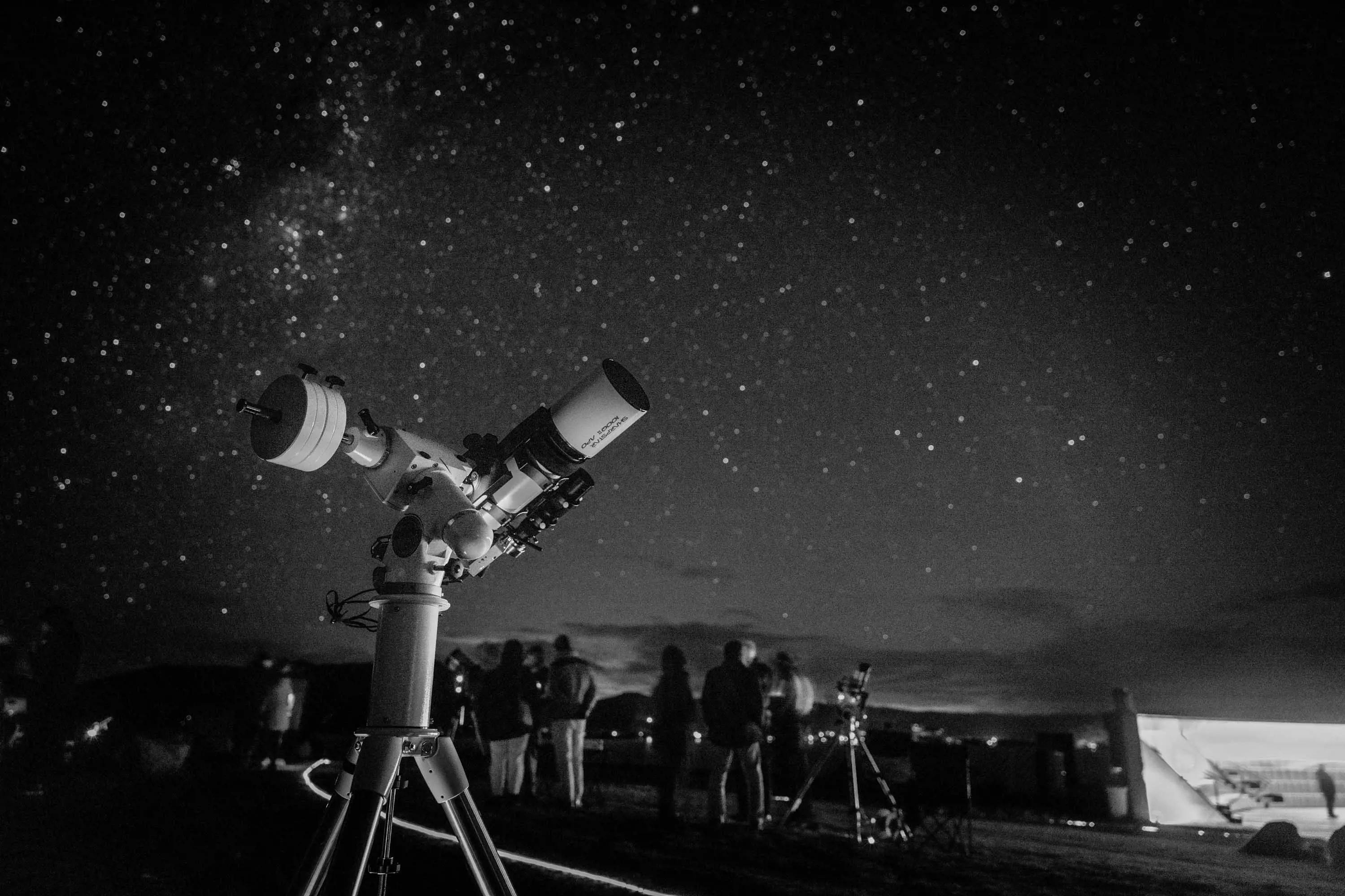 A large telescope stands in the foreground, while people stand in a group, gazing at the clear night sky filled with stars.