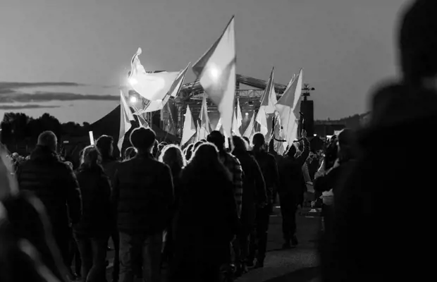 A large procession of people carrying flags walk in a long, sprawling line towards a performance stage in the background.