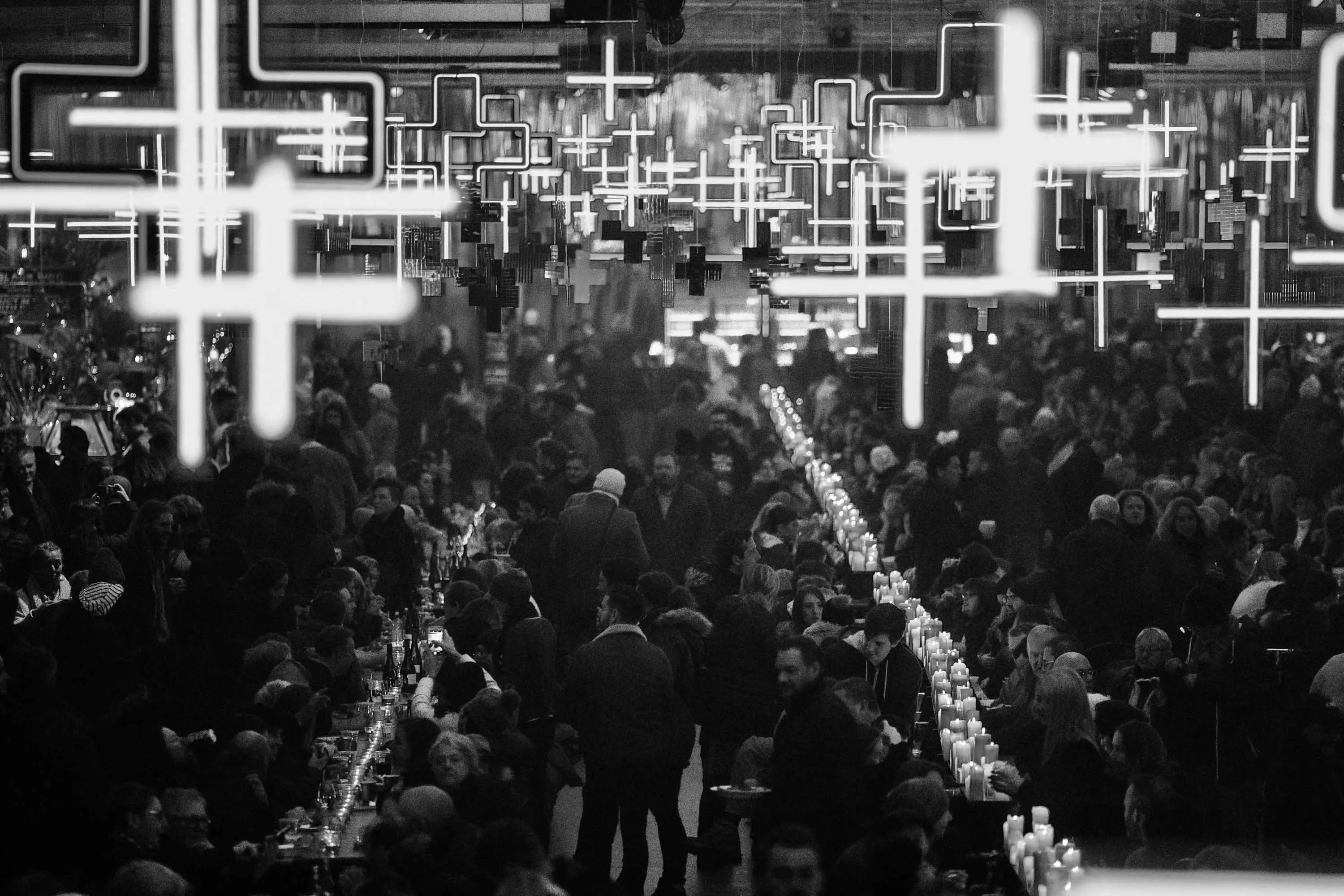 A large converted pier shed is lined with long tables. Thousands of people sit along them, eating food to candlelight. Hundreds of large cross shaped lights are suspended overhead.
