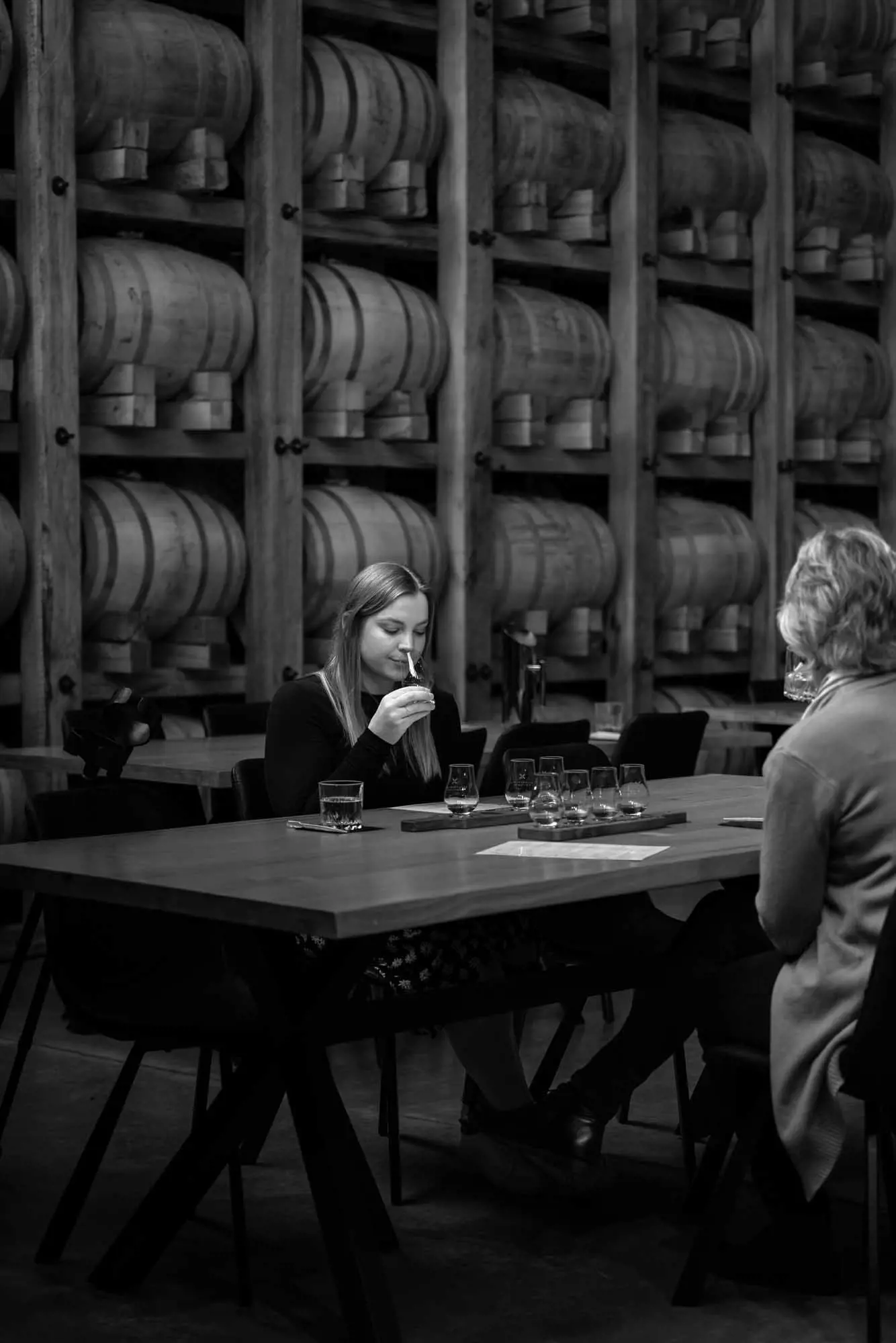 A young woman and an older woman sit at a large wooden table and taste spirits with a wall filled with barrels in the backgound.