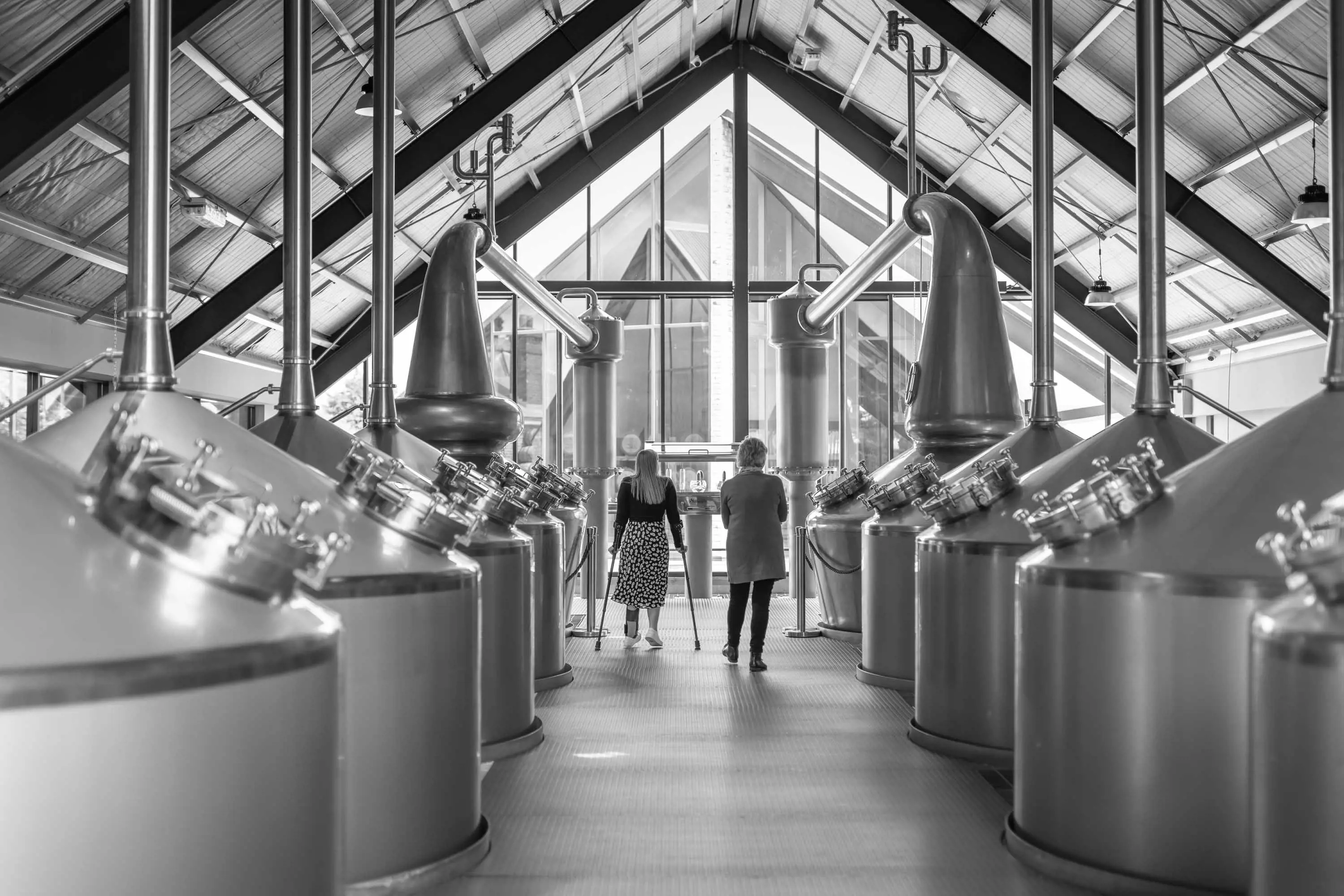 Two people walk between several large distillers housed in a triangular, tall-roofed industrial shed.