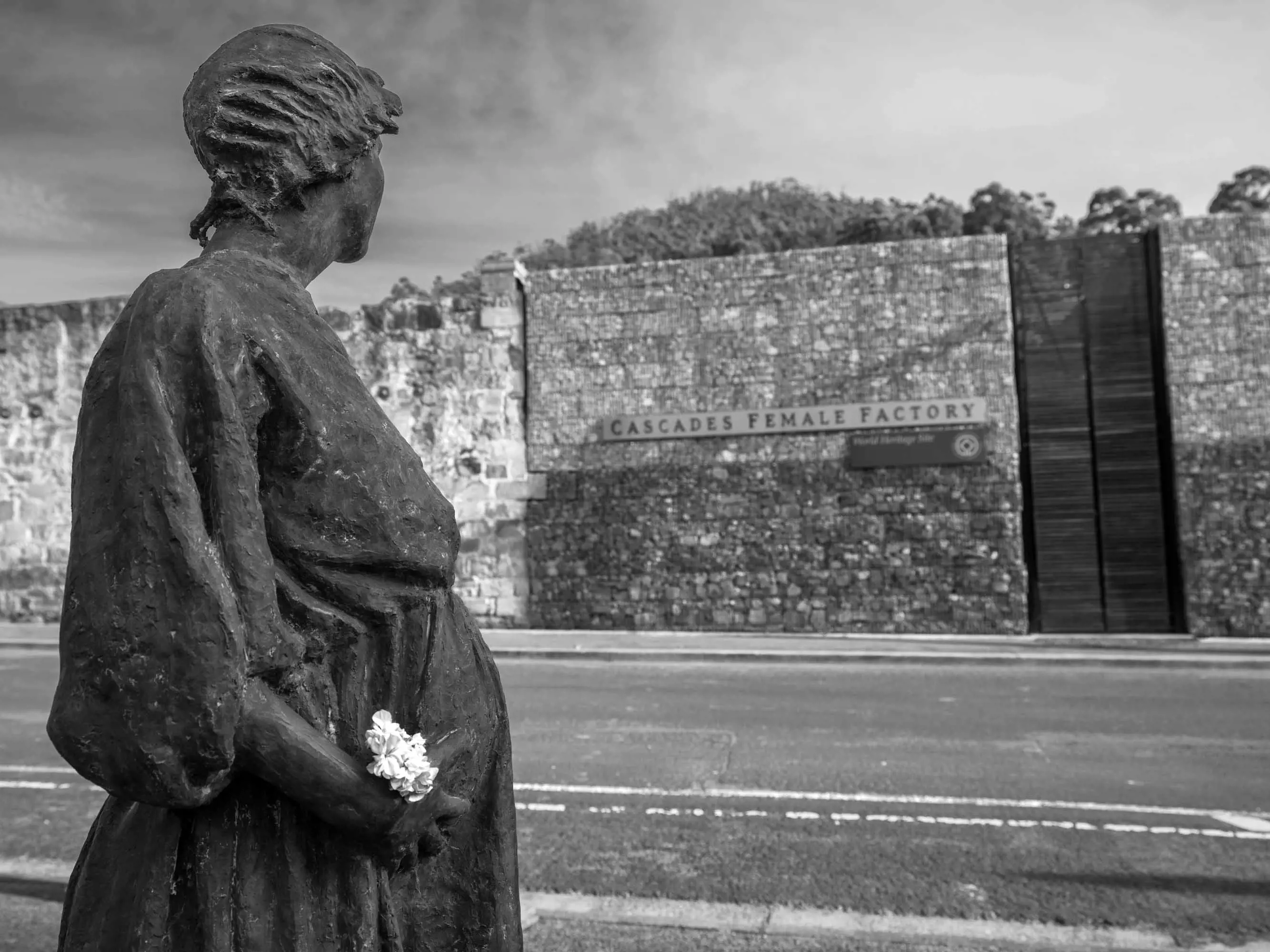 A bronze statue of a woman in early 19th century clothing stands on the side of a road opposite the large stone wall entrance to a historic site.