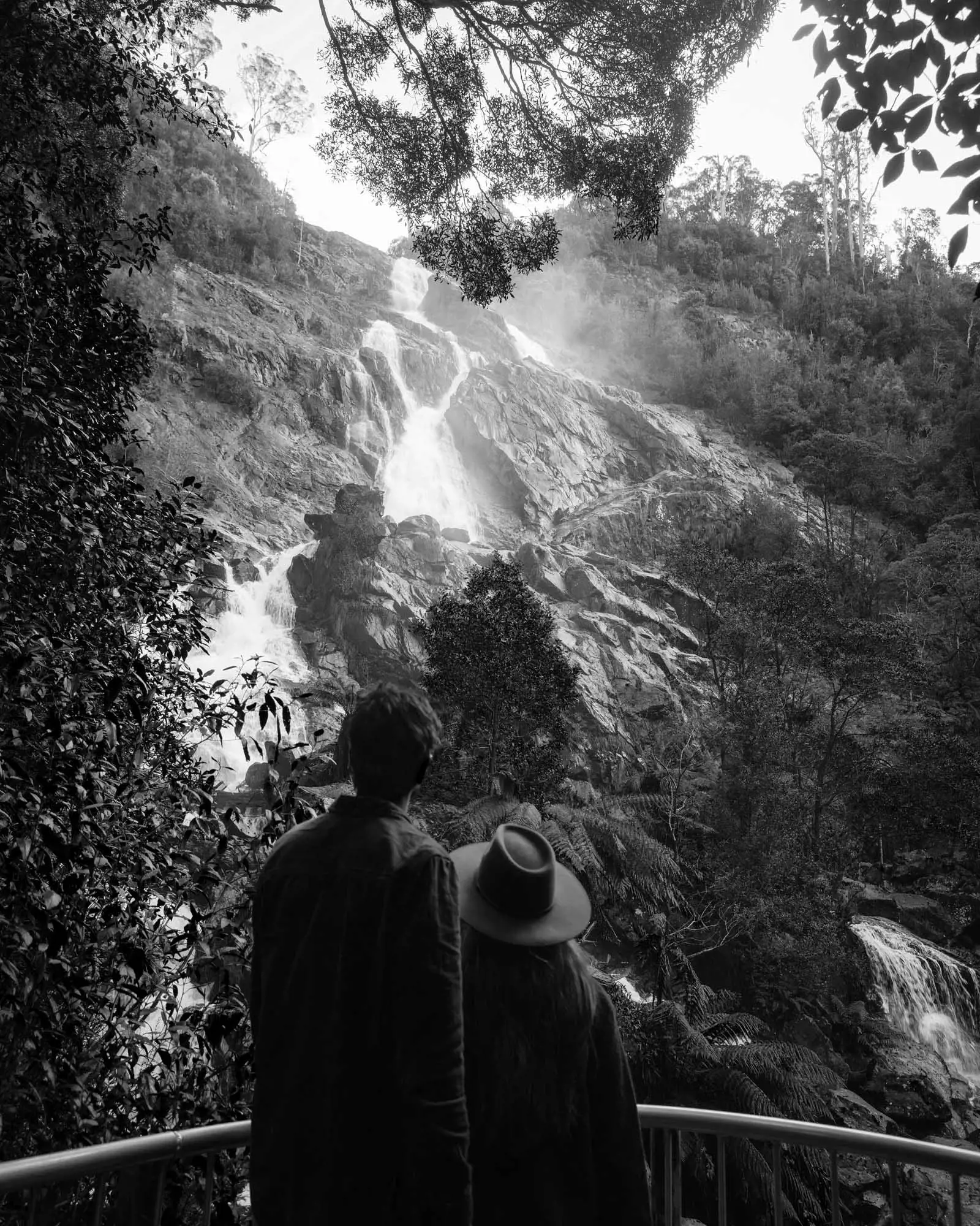 A couple stand at the base of a tall waterfall, that spills over the rocky outcrops of a cliff face above them.