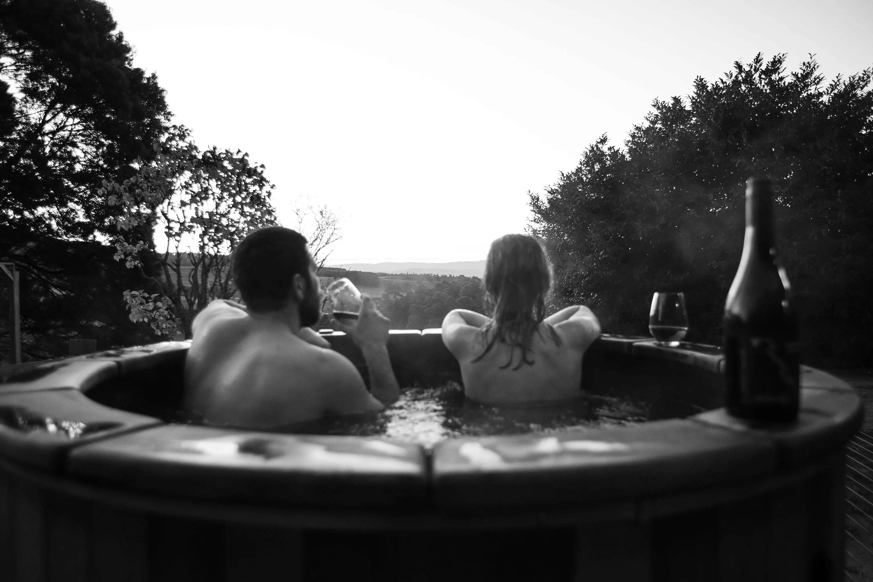 Couple in an outdoor wooden hot tub, drinking wine and taking in the view.