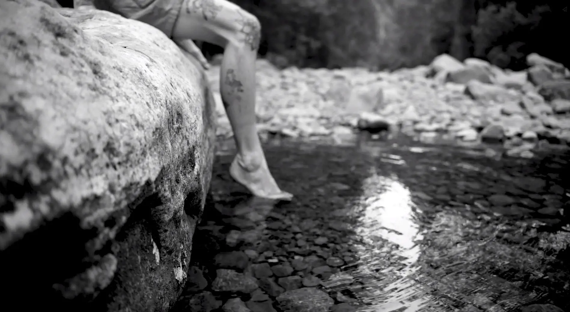 A man with tatooed legs sits on a large rock and dips his toe in cold, clear water.