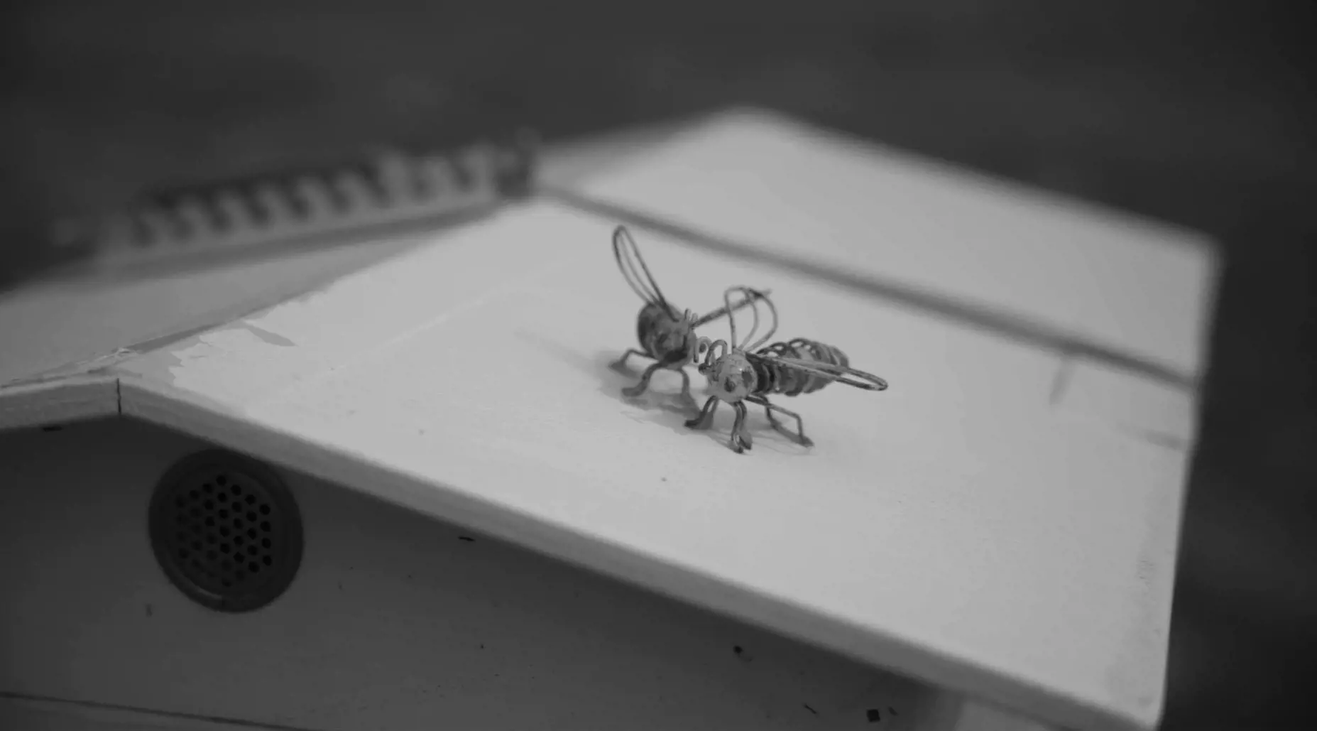 A pair of ornamental bees made of wire sit on top of the roof of a white beehive.