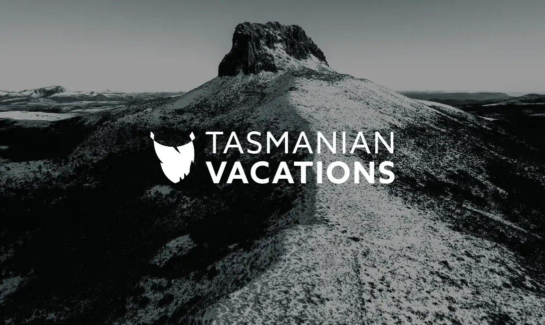 A monochrome photo of a mountain dusted with snow. It is overlaid with text saying "Tasmanian Vacations" and an icon of Tasmania.
