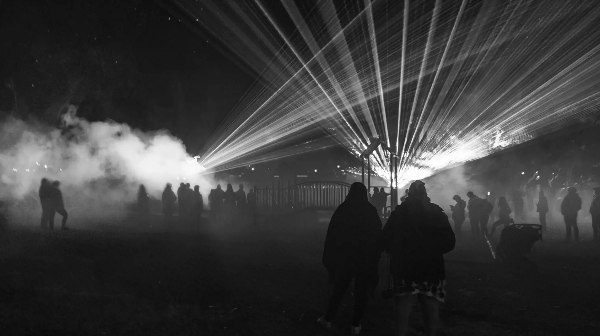 Groups of people stand in a park and watch a laser show illuminate the mist from a smoke machine.