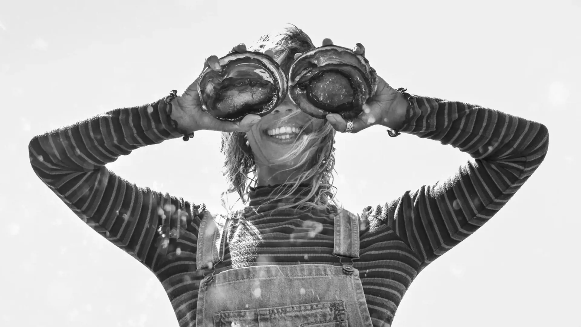 A woman holding two large abalone shells in front of her eyes, playfully pretending they are her eyes