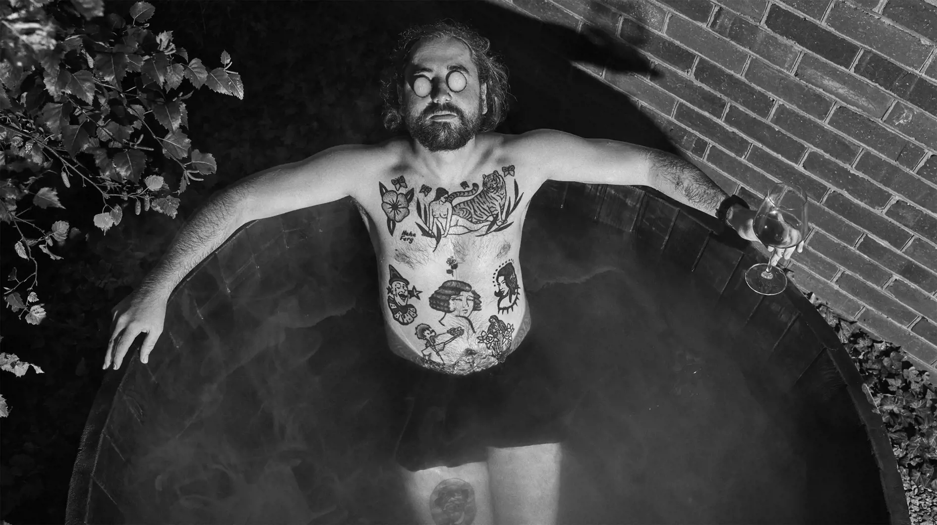 Tattooed man relaxing in wooden bath tub with a glass of wine and cucumbers over his eyes.