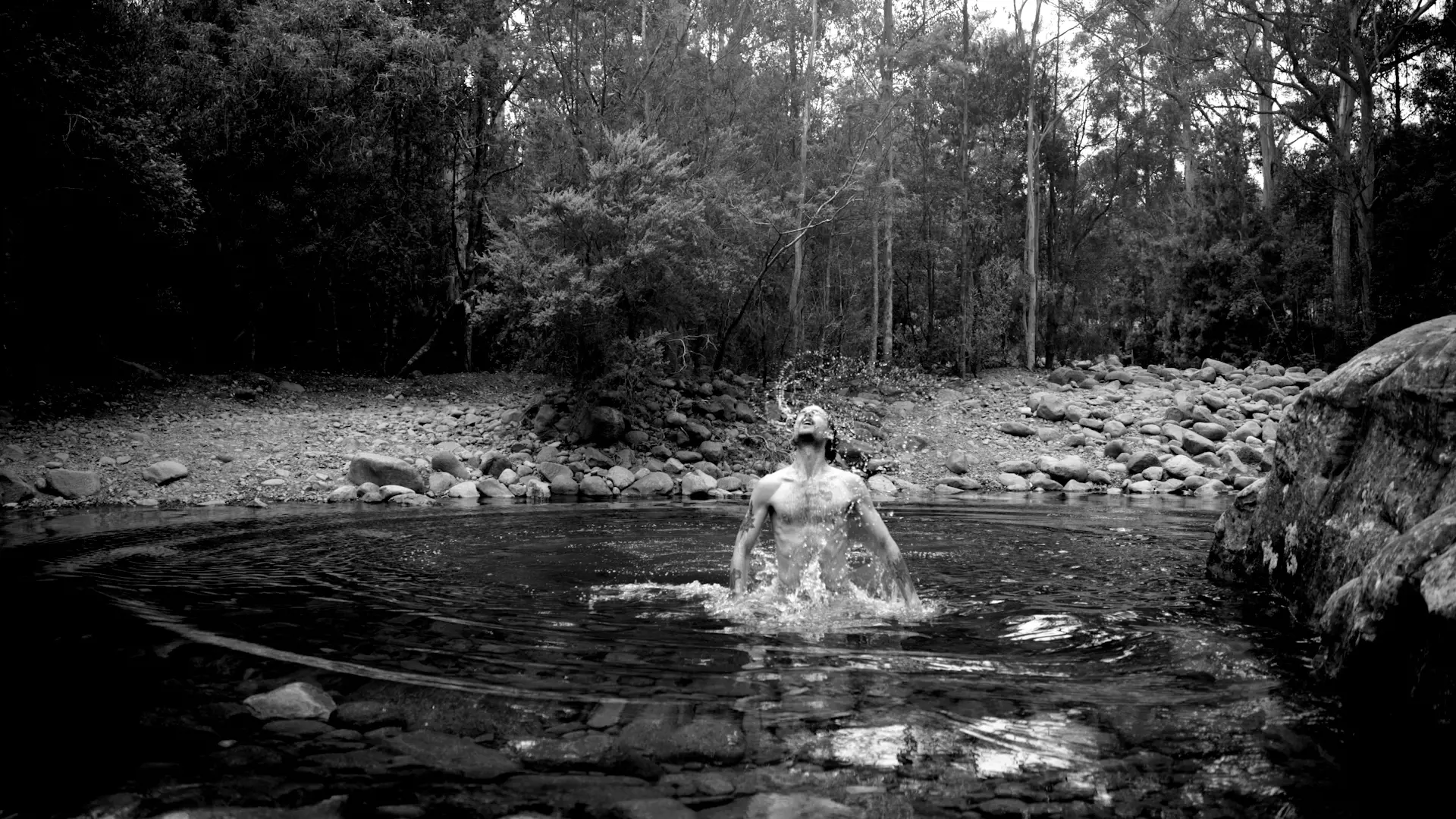 A man jumps up from beneath the surface of a small, rocky pond situated within a forest, splashing water.
