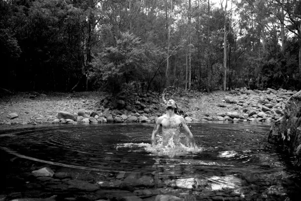 A man jumps up from beneath the surface of a small, rocky pond situated within a forest, splashing water.